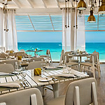 Elegant beachfront dining area perfect for a destination wedding, with set tables, white chairs, and a view of a turquoise sea through large windows.