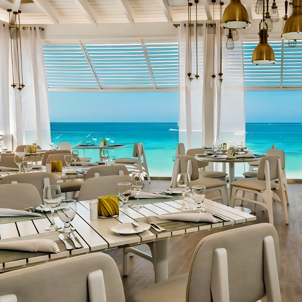 Elegant beachfront dining area perfect for a destination wedding, with set tables, white chairs, and a view of a turquoise sea through large windows.