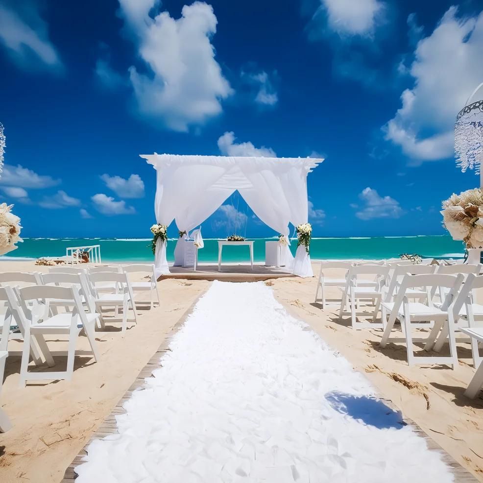 Beach wedding setup with white chairs, an aisle on sand, and a white canopy overlooking turquoise ocean and a blue sky with clouds.