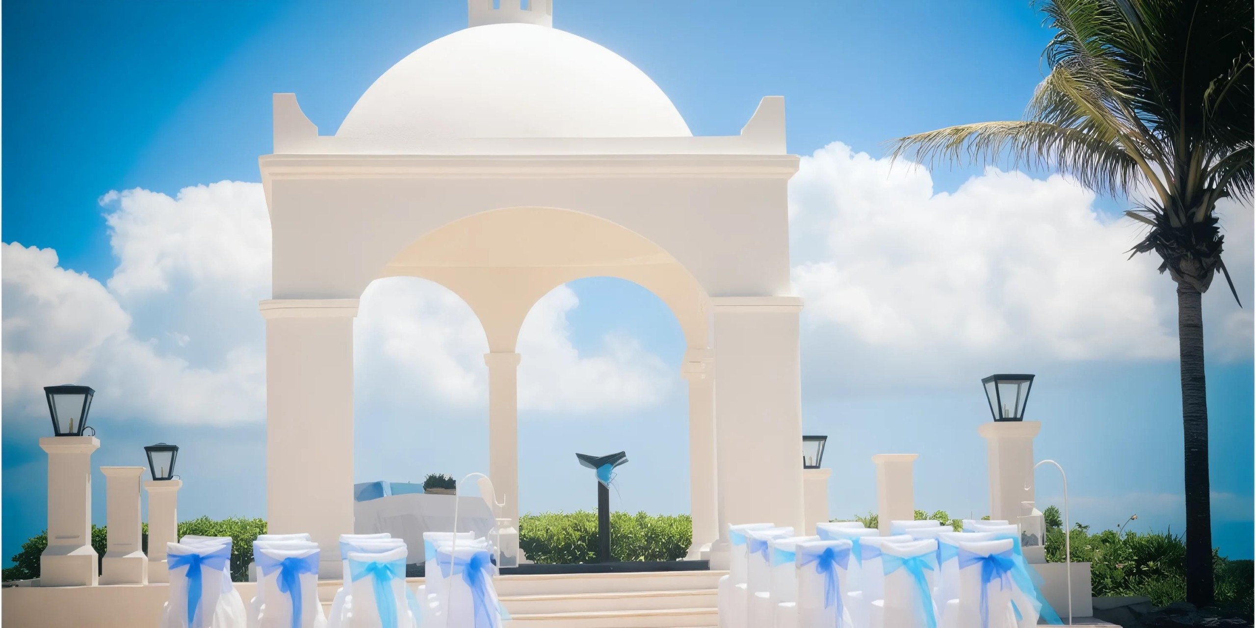 An outdoor wedding venue with a white gazebo, decorated chairs, and a palm tree under a clear blue sky.