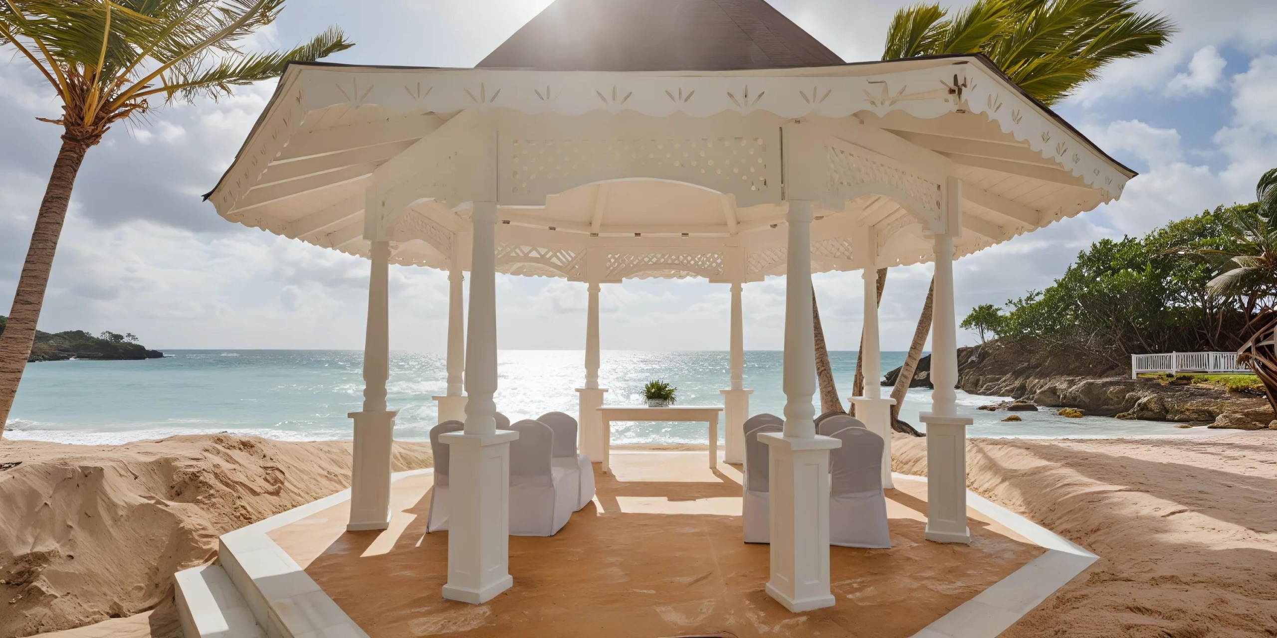 White gazebo with seating on a sandy beach, overlooking the ocean under a partly cloudy sky.