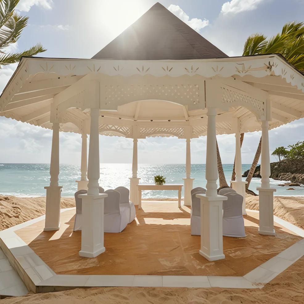 A white gazebo on a sandy beach with palm trees, overlooking the ocean under a partly cloudy sky.