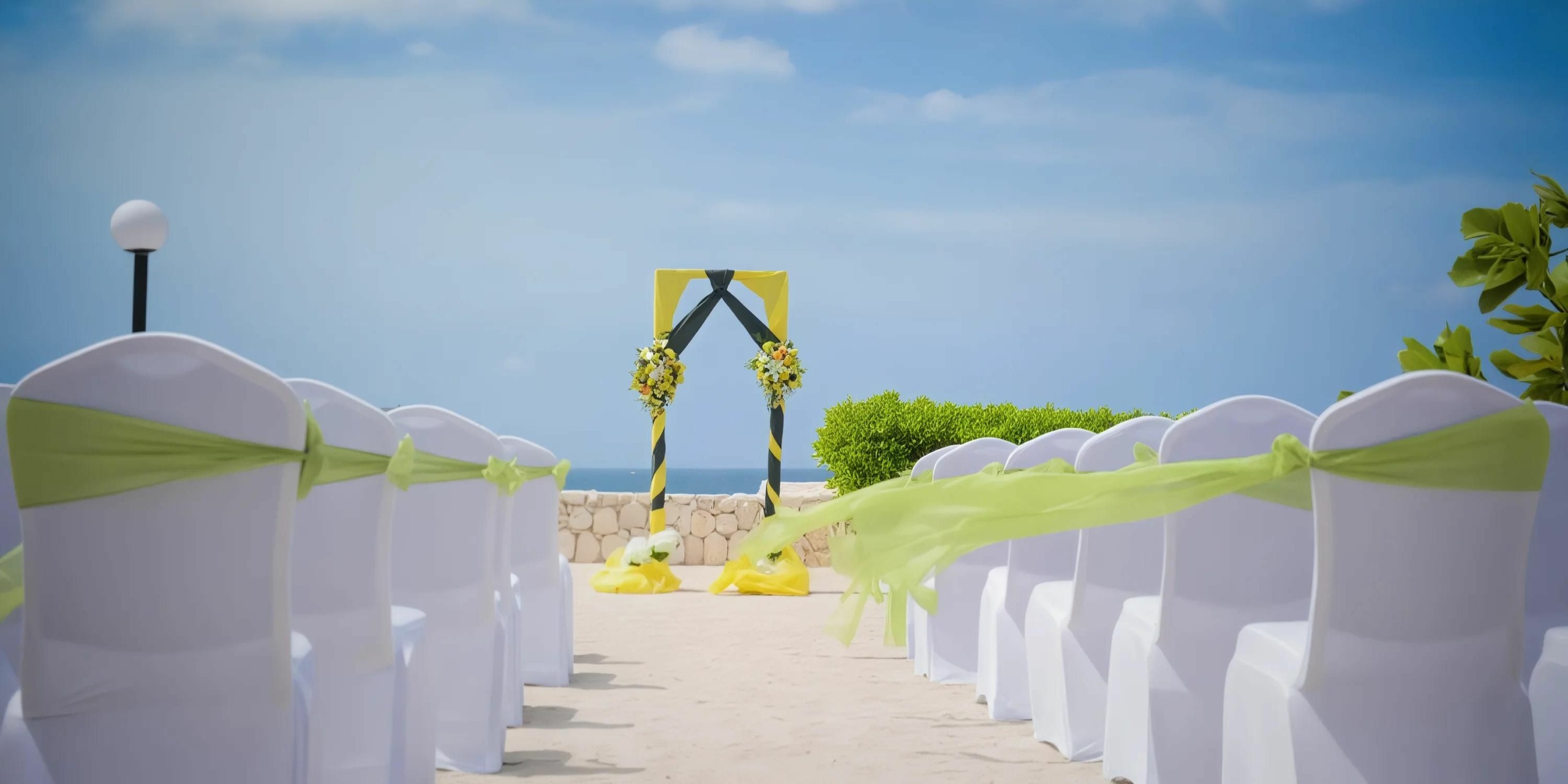 Outdoor beach wedding ceremony setup with white chairs adorned with green sashes facing a decorated arch. Blue sky and ocean visible in the background.
