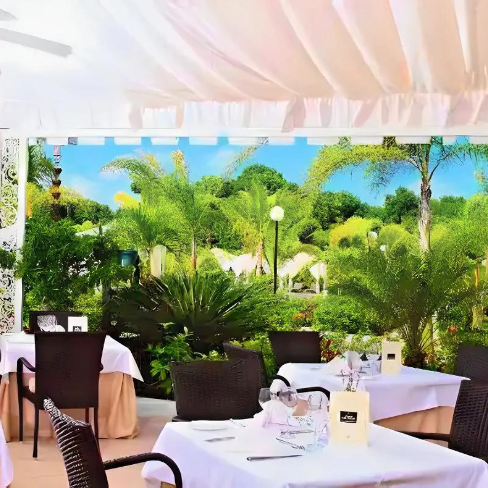 Outdoor restaurant terrace ideal for destination weddings, with white draped textiles, neatly set tables, and a lush green garden view under a bright sky.