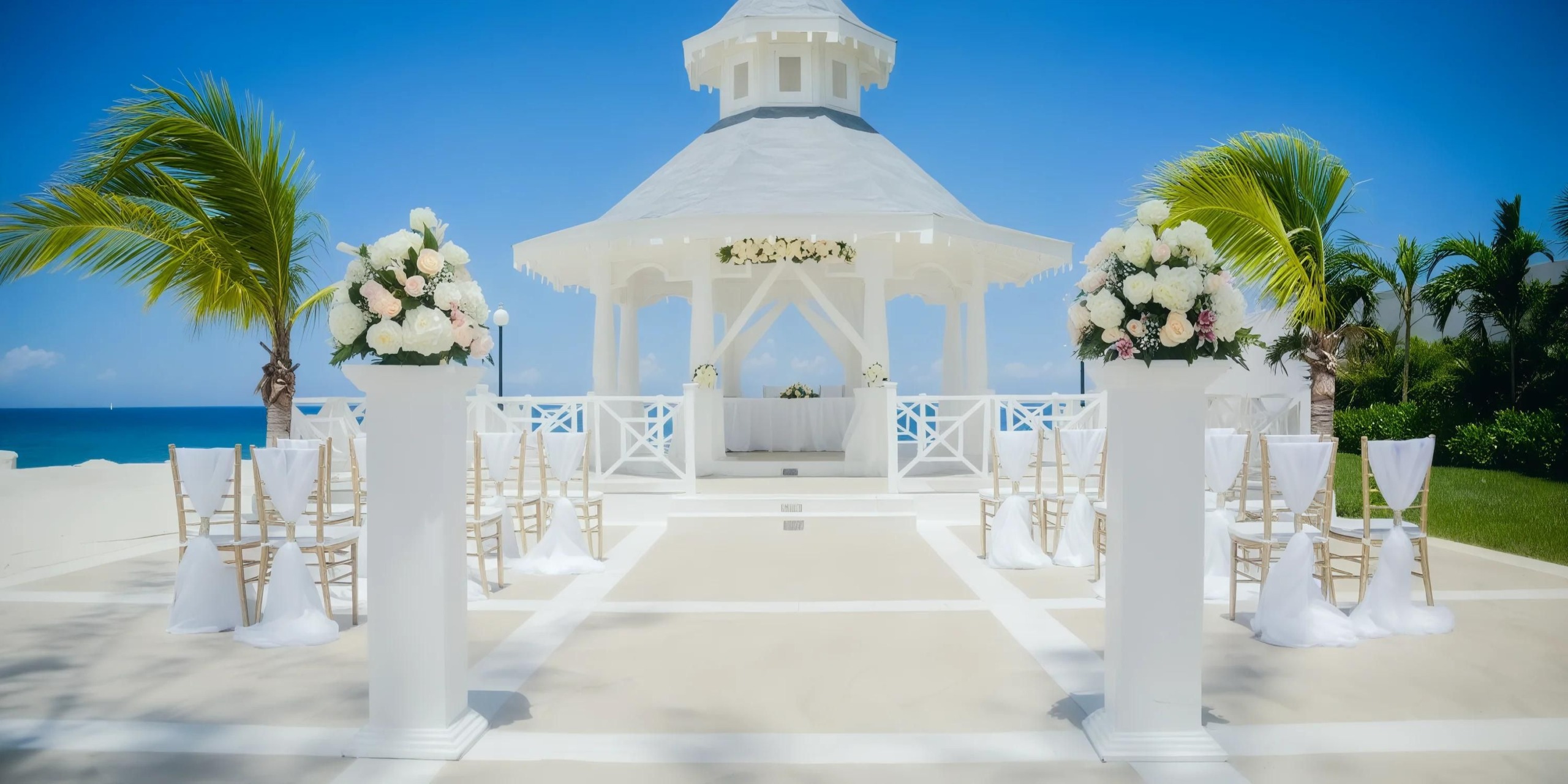 Imagine an outdoor wedding setup at the Bahia Principe Luxury Runaway Bay, with a pristine white gazebo, elegant floral arrangements, and rows of chairs set under a clear blue sky by the beach.