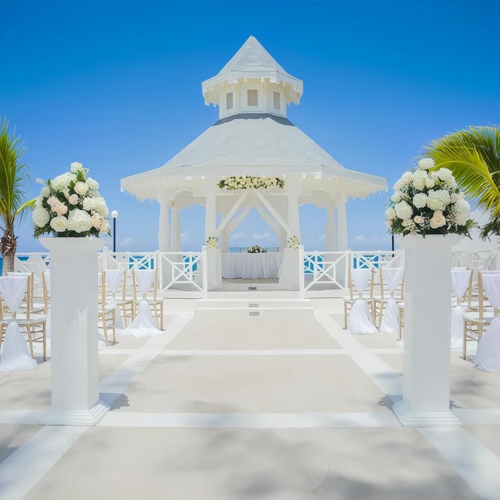 White wedding gazebo by the sea with flower arrangements and chairs in an outdoor setting under a clear blue sky.