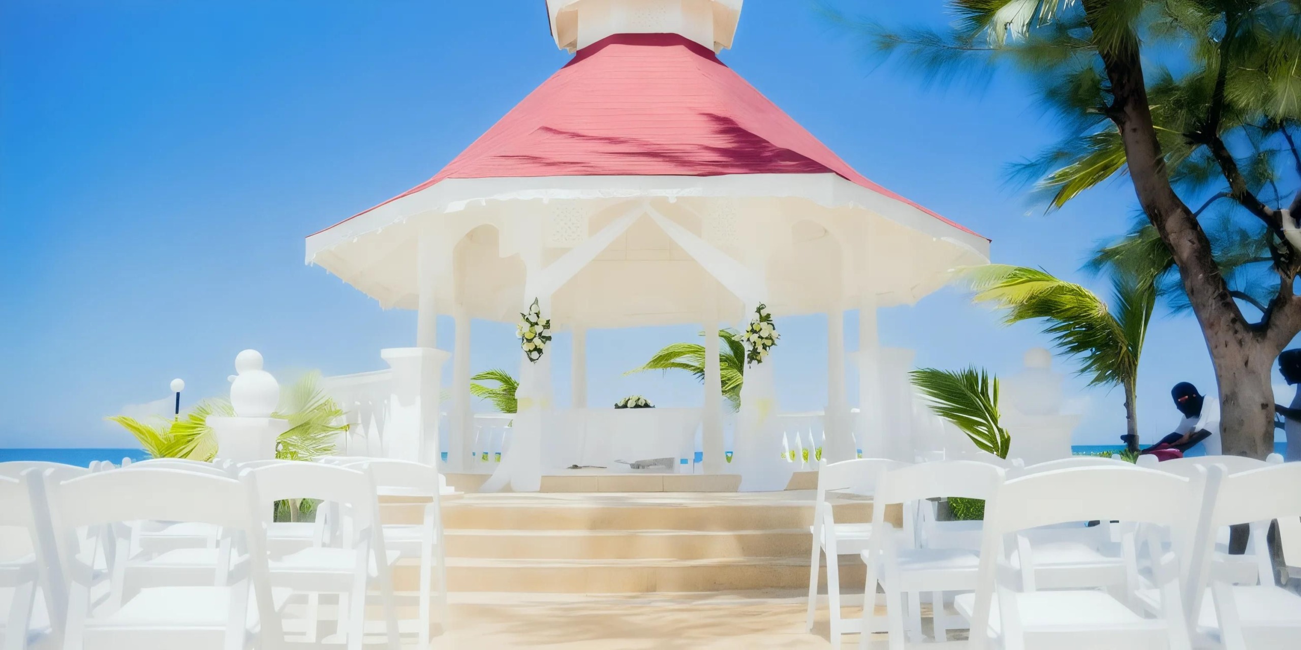 A white gazebo with a red roof is set for an outdoor event, surrounded by palm trees and white chairs, under a clear blue sky.