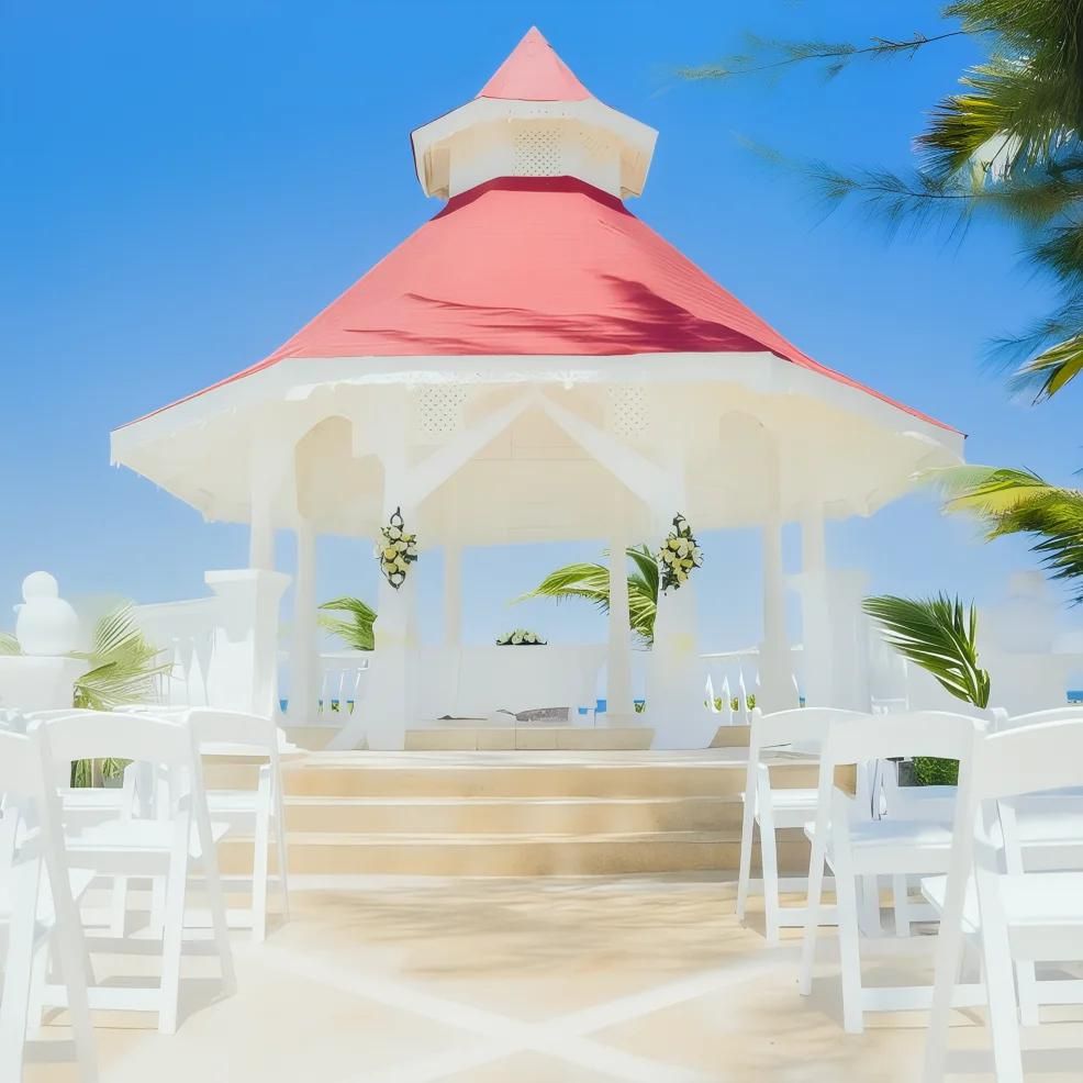 A white gazebo with a red roof is set up for an event, surrounded by white chairs, against a backdrop of blue sky and palm trees.