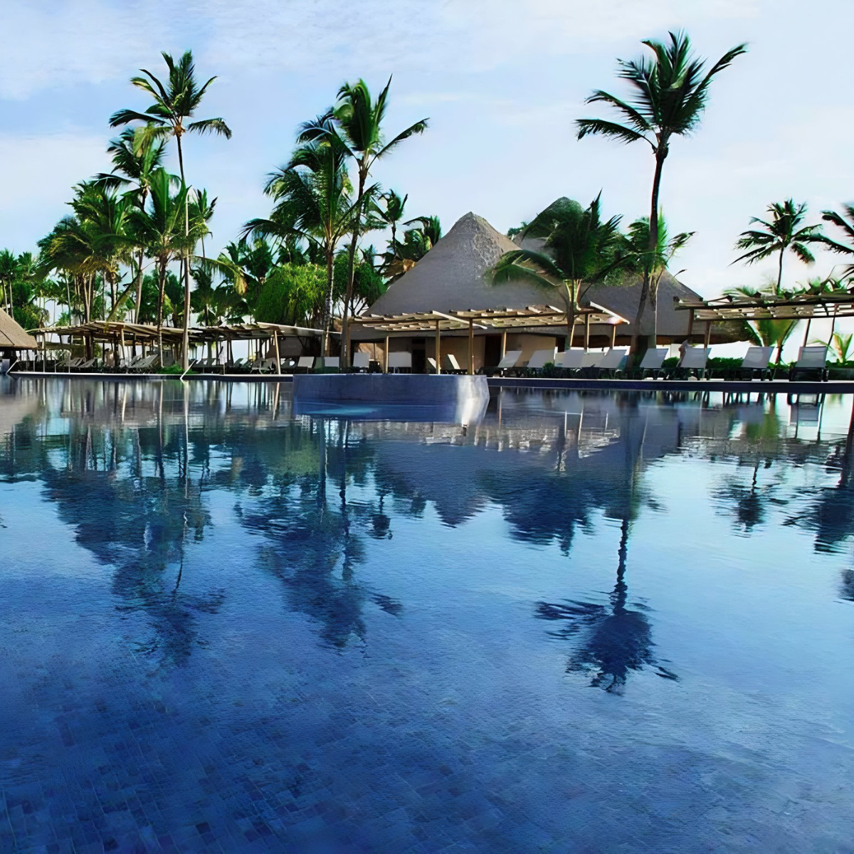 A tranquil Barcelo Bavaro Palace destination wedding resort pool surrounded by palm trees with thatched-roof bungalows reflected in the water under a clear sky.