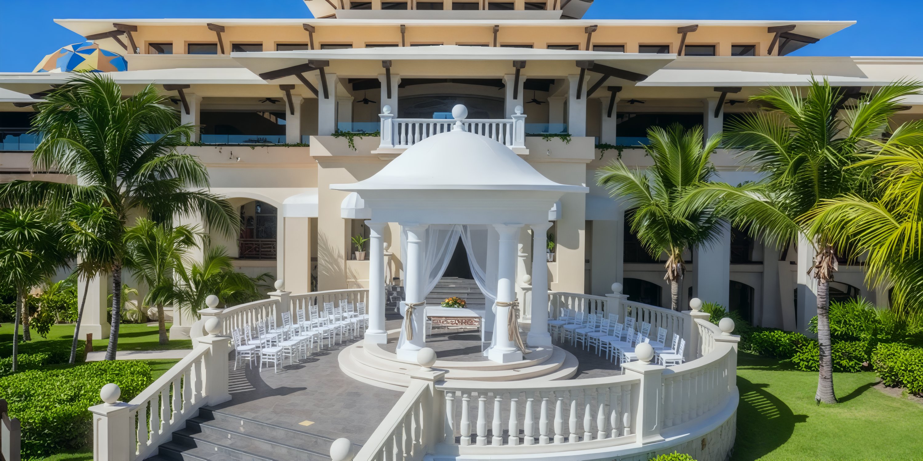 Barcelo Maya Tropical offers a picturesque setting for a destination wedding, featuring a white gazebo with seating, surrounded by swaying palms and adjacent to a grand building.