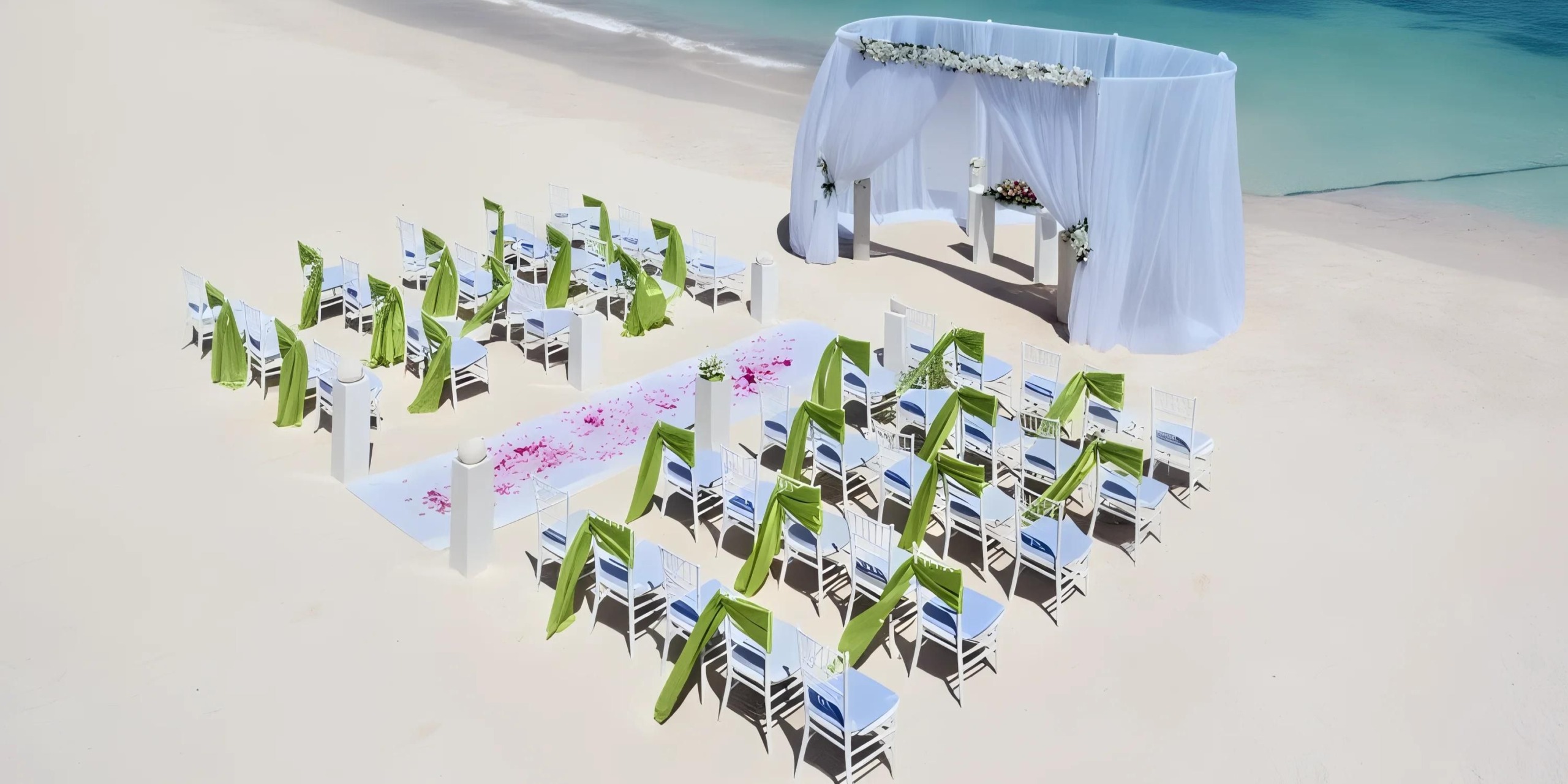 The Barcelo Maya Tropical offers a beach wedding setup featuring white chairs adorned with green sashes, all arranged to face a beautifully decorated archway underneath a canopy by the ocean. The sandy aisle is elegantly lined with pink petals.