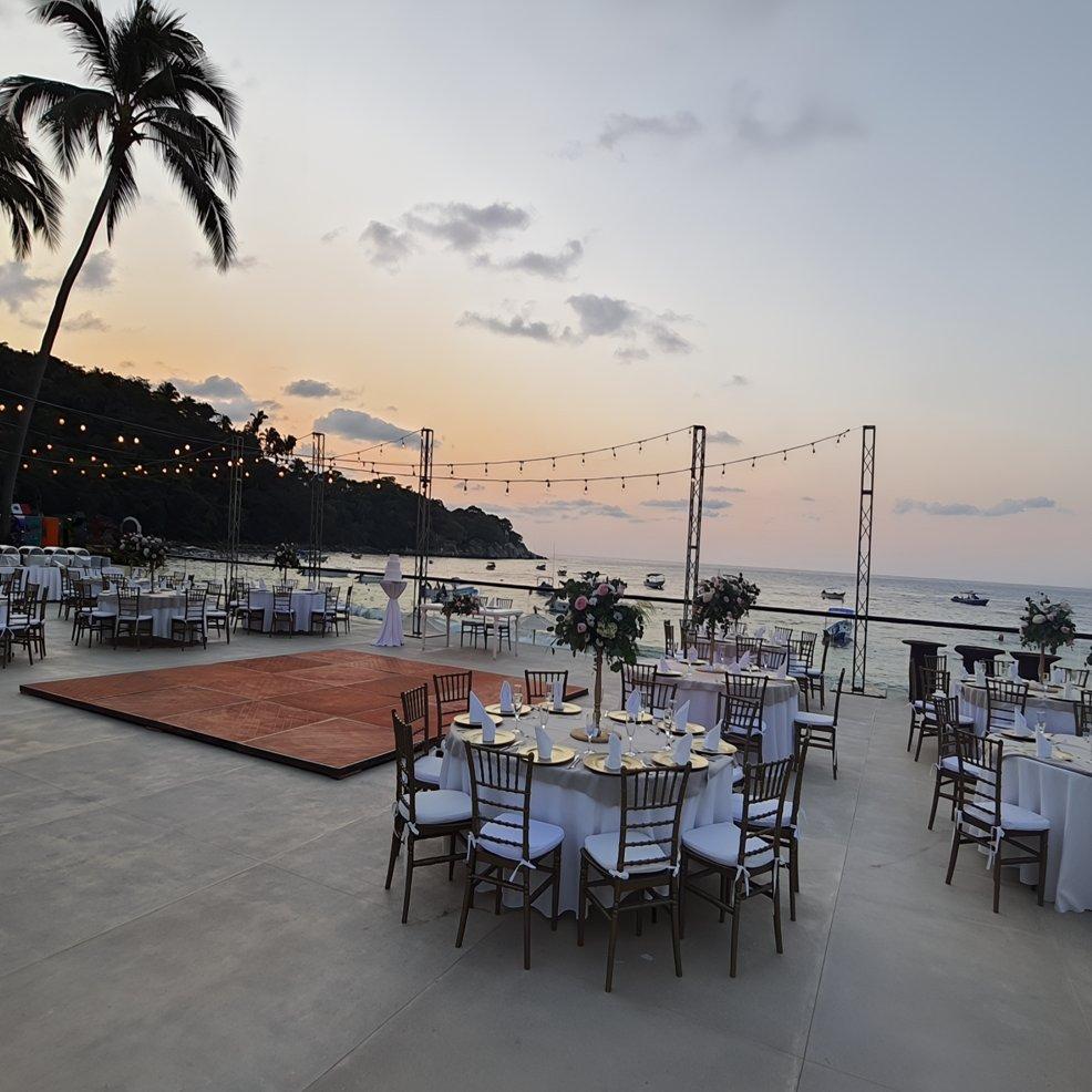 Outdoor beachfront destination wedding setup with round tables, chairs, and string lights at sunset.