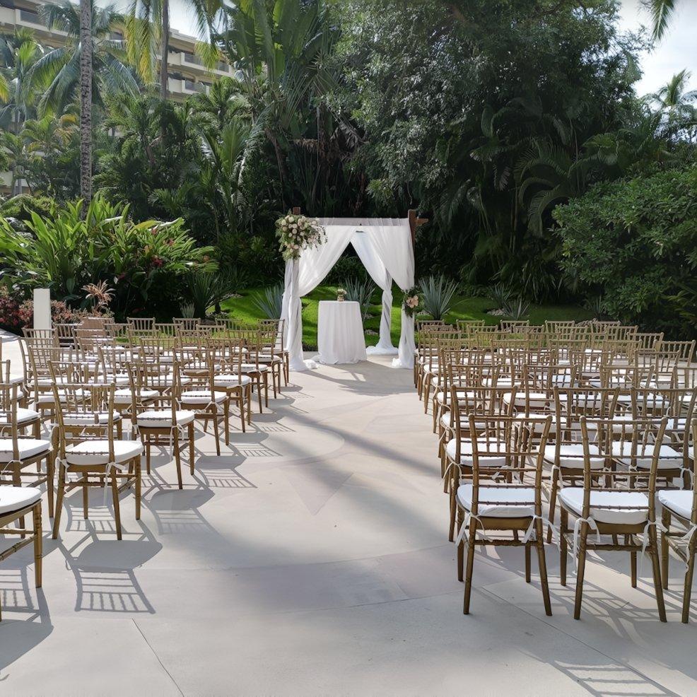 Destination wedding setup outdoors with rows of chairs arranged toward a white, floral-decorated arch.