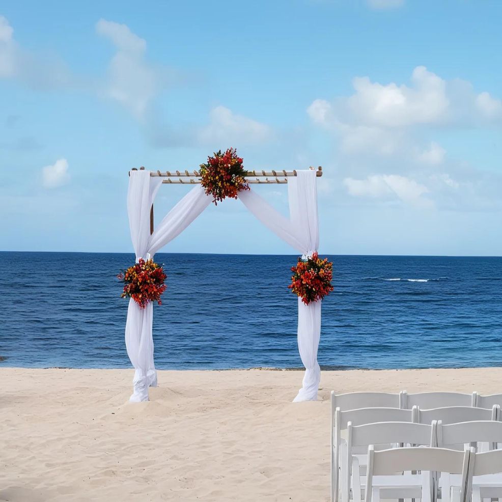 A decorated wedding arch with white fabric and red floral arrangements on a sandy beach with the ocean in the background. White chairs are arranged in rows on the right side of the image.