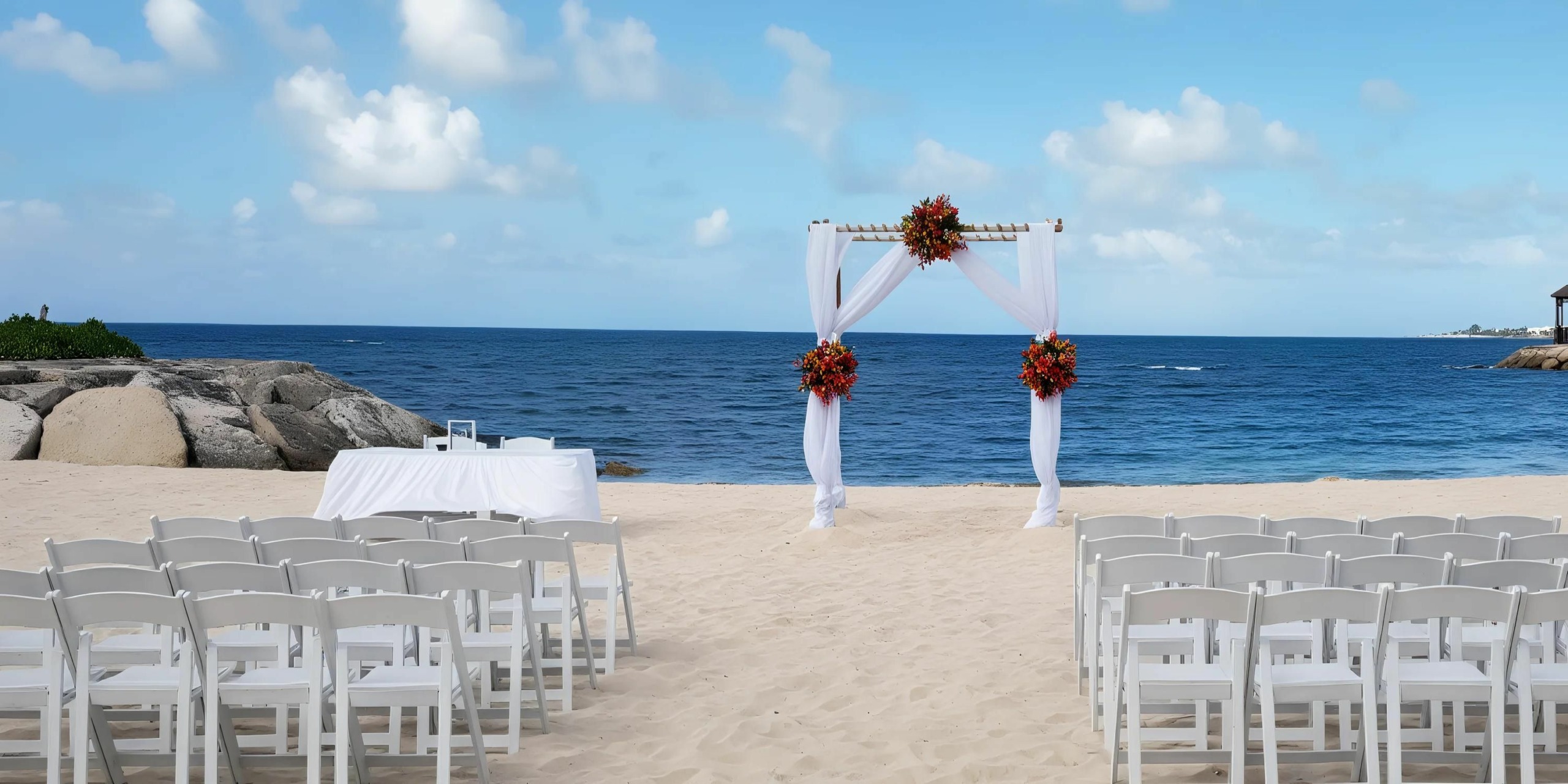 A beach wedding setup with white chairs arranged in rows facing an arch decorated with red flowers and white fabric, overlooking the ocean on a sunny day.