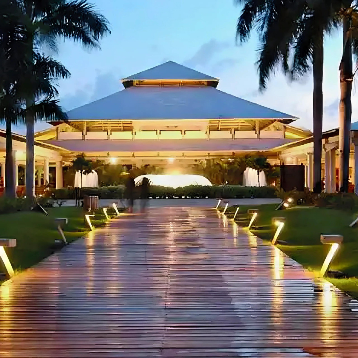 A lit wooden walkway leading to Catalonia Bavaro Beach, a large, illuminated building with a pyramid-shaped roof at twilight, flanked by palm trees and grass, perfect for a destination wedding.