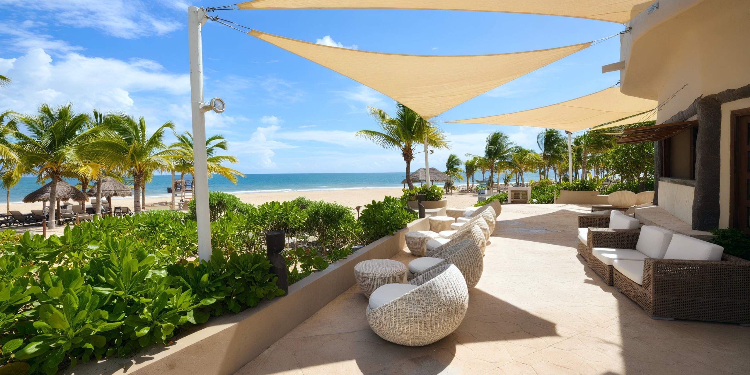 Experience the stunning Catalonia Playa Maroma with its beachside patio featuring white chairs and tables beneath triangular shade sails. This idyllic setting is surrounded by lush greenery and palm trees, offering breathtaking views of a sandy beach and the mesmerizing blue ocean.
