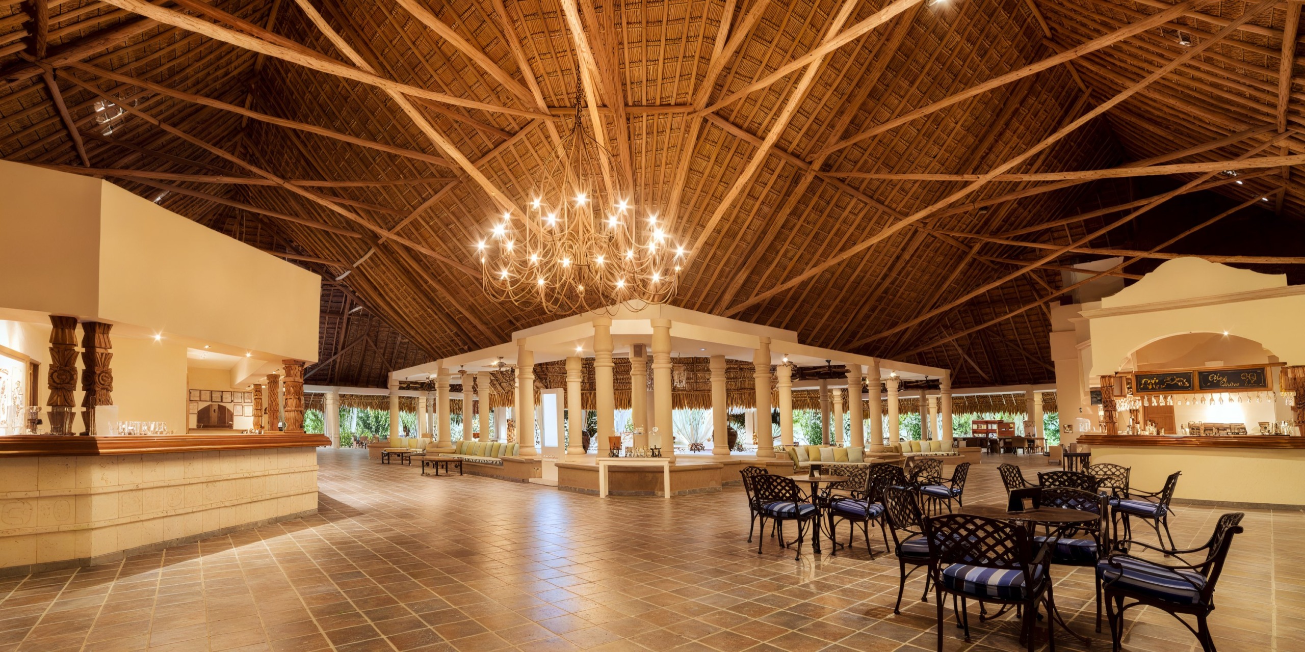 The Catalonia Playa Maroma features a spacious restaurant interior with a high thatched ceiling, chandelier lighting, and a tiled floor. Tables and chairs are arranged under wooden beams, creating a rustic and open atmosphere.