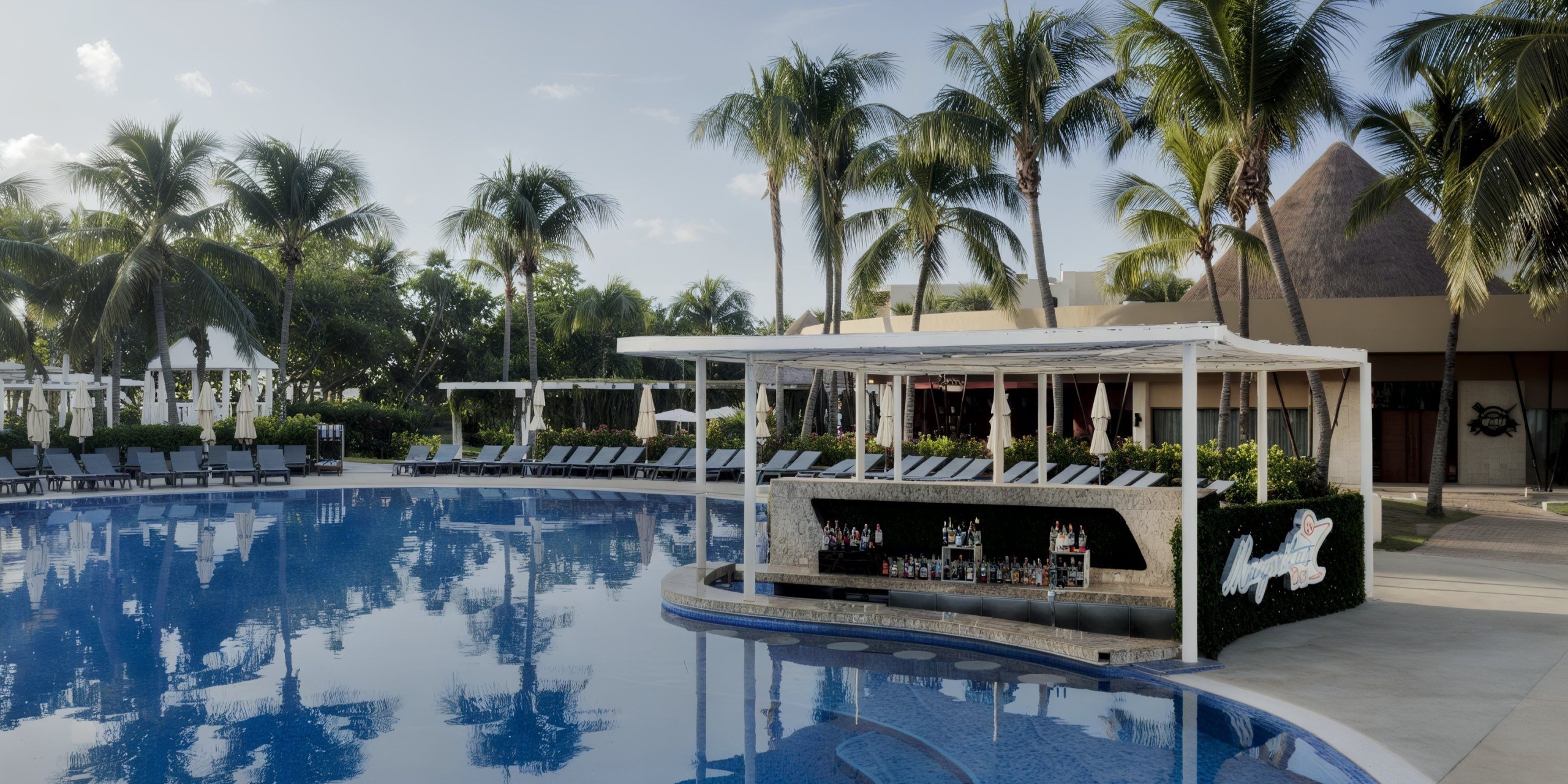 Experience the Catalonia Playa Maroma, featuring a tropical resort pool adorned with palm trees, lounge chairs, and a swim-up bar under a canopy. In the background, you'll find a large building with an iconic thatched roof.