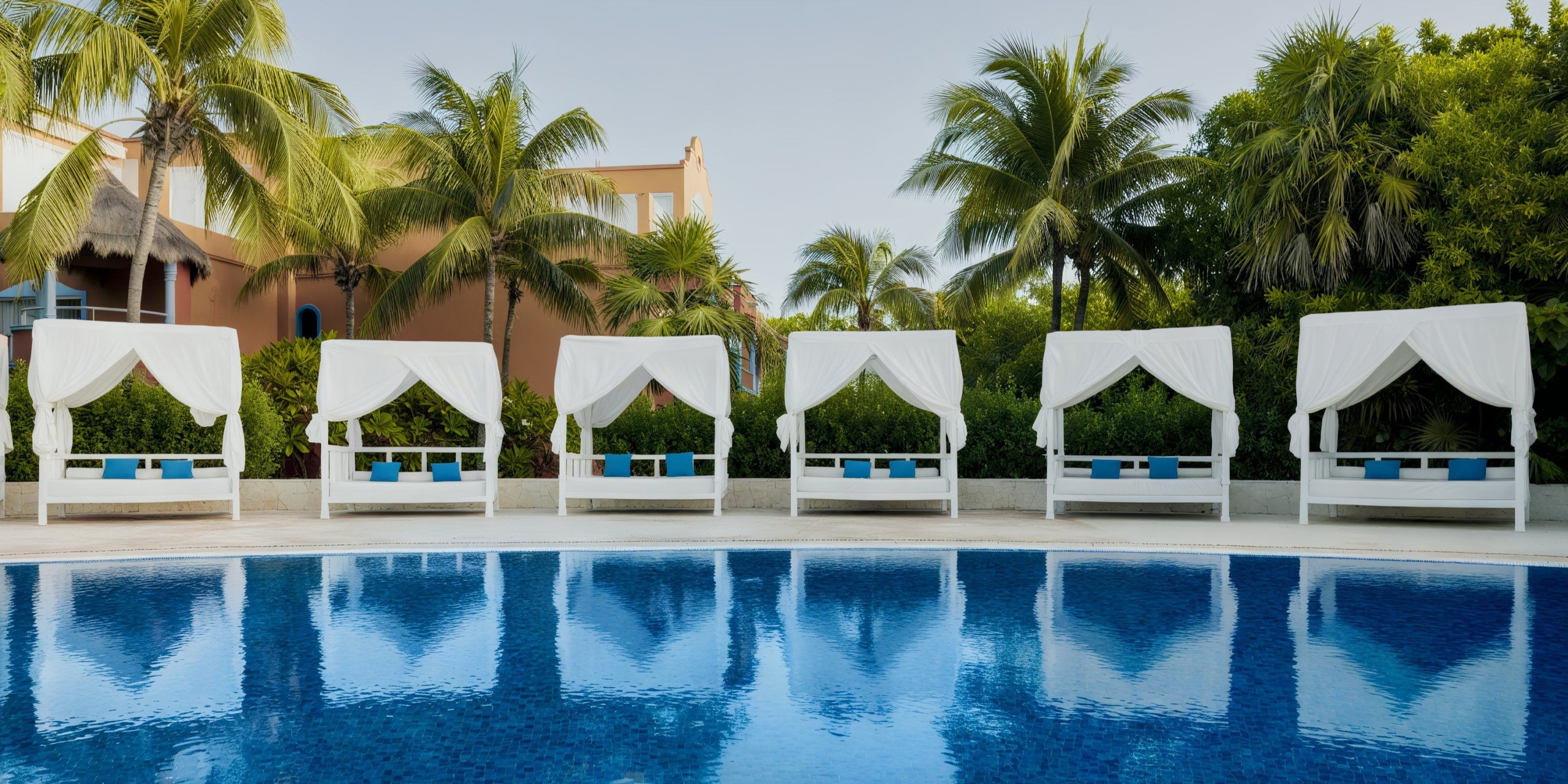 A view of the Catalonia Playa Maroma showcases six white cabanas, each adorned with blue cushions, set against a backdrop of palm trees and a beige building.