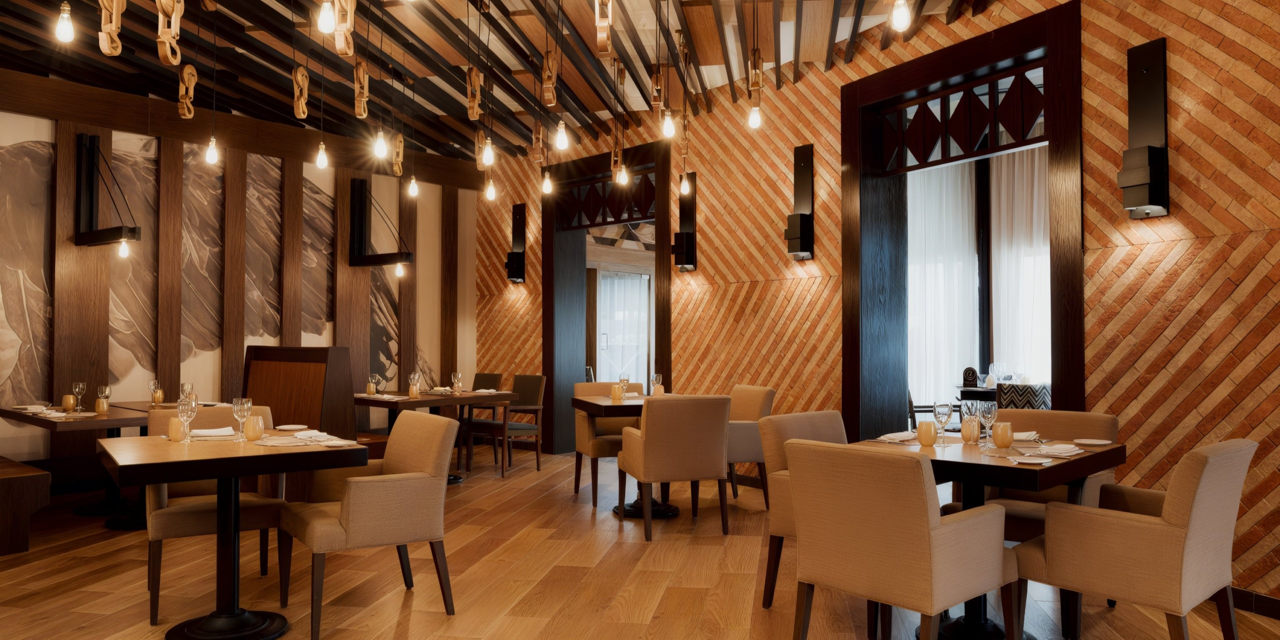 The Catalonia Playa Maroma restaurant interior features wooden decor, beige chairs, dark tables, and ceiling lights. The walls showcase artistic paneling with striped patterns. The room is currently unoccupied.