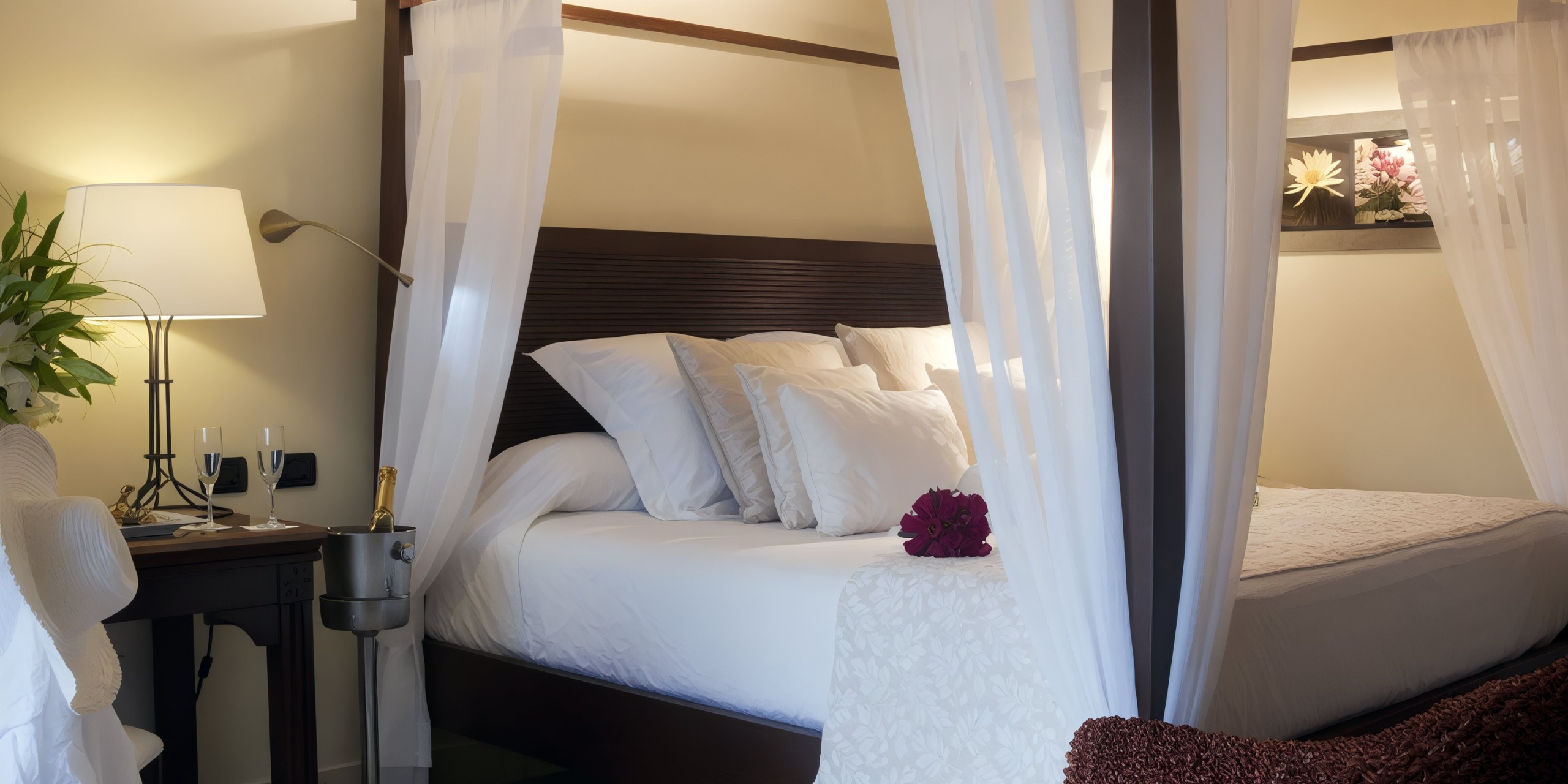 A cozy bedroom at the Catalonia Playa Maroma features a four-poster bed draped with white curtains and pillows, accompanied by a lamp and a table holding champagne on the side. A floral artwork adorns the wall.
