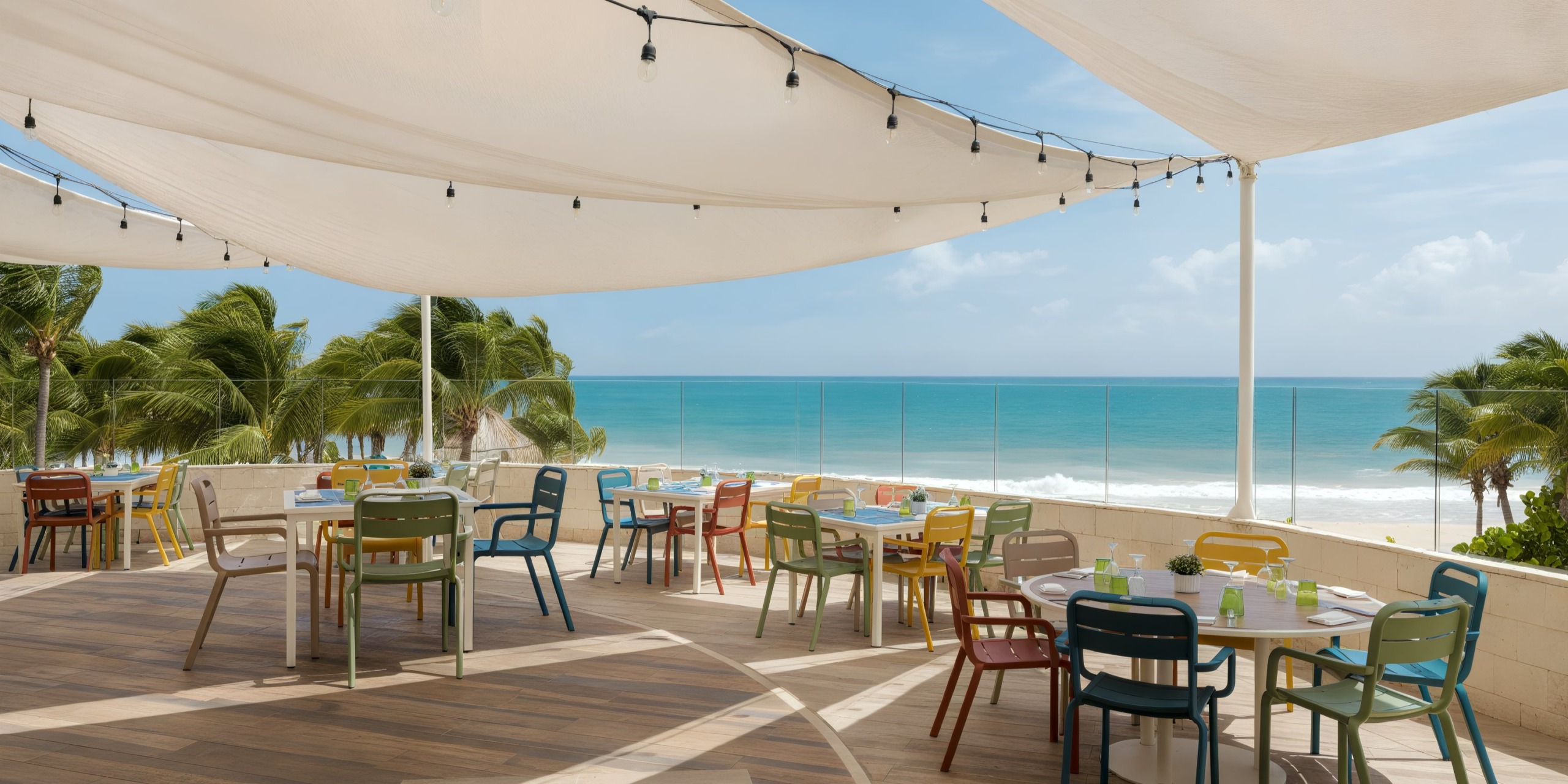 Rooftop patio at Catalonia Playa Maroma featuring colorful chairs and tables beneath white canopies, offering a view of the beach with palm trees and the ocean.