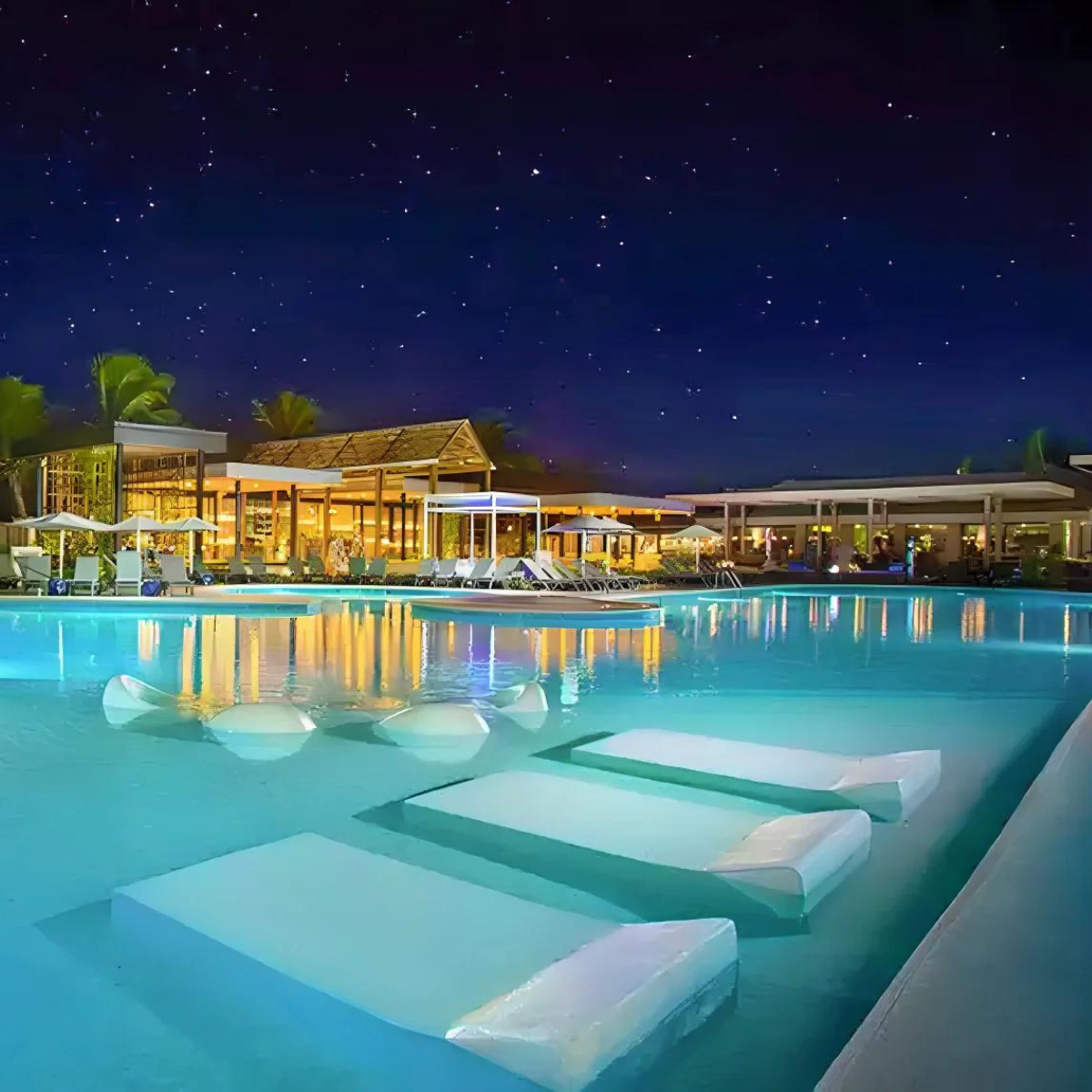 A Catalonia Royal Bavaro destination wedding resort pool illuminated at night with floating sunbeds, a starry sky above, and a lit-up building in the background.