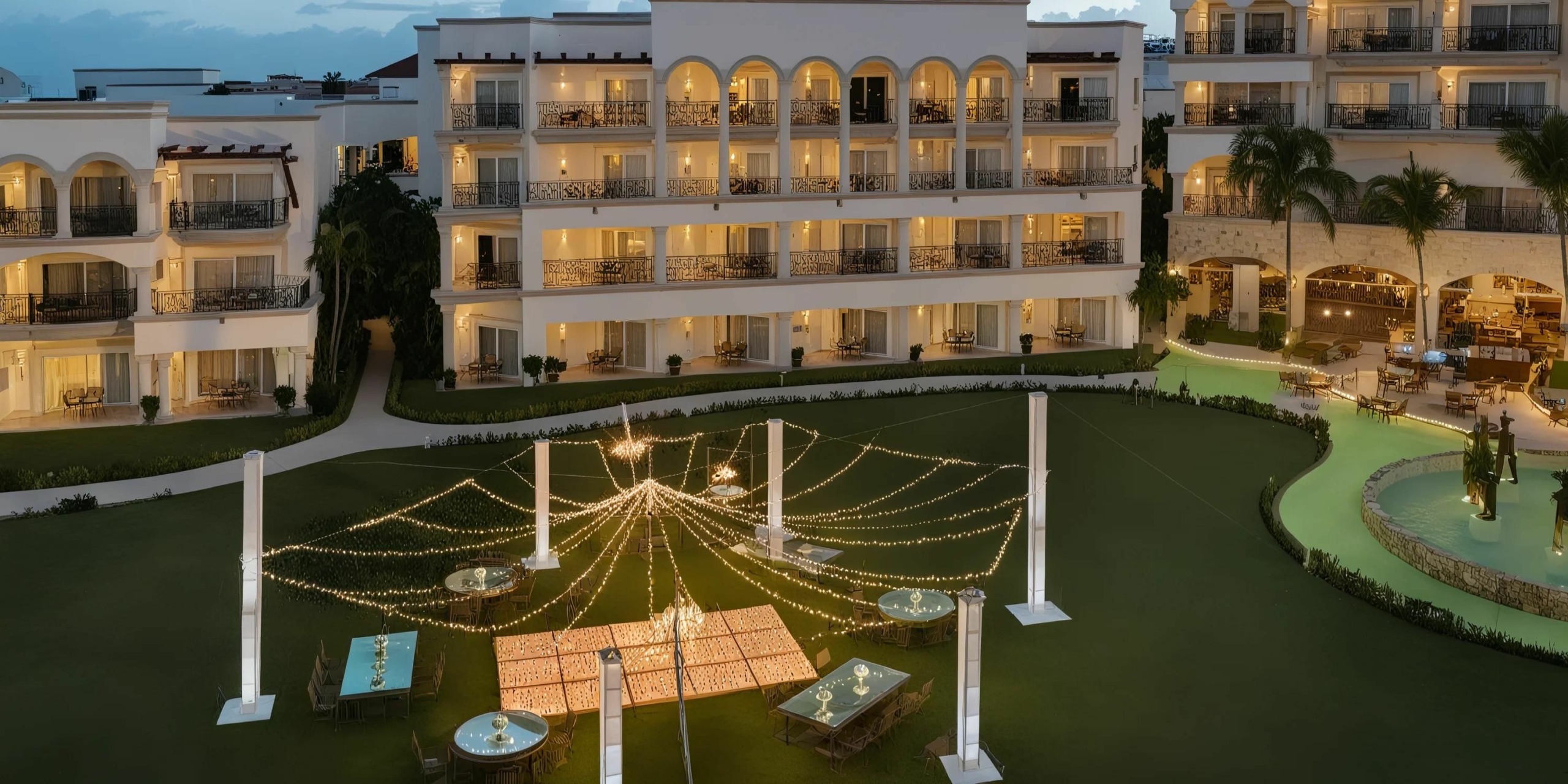 An outdoor event setup with string lights and tables on the lawn in front of the Hilton Playa del Carmen, a multi-story building featuring illuminated balconies.