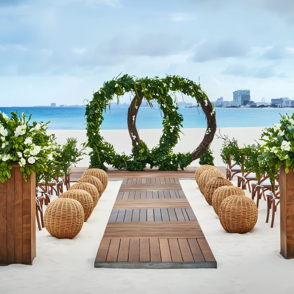Beach wedding setup with a wooden aisle, wicker chairs, and a decorative greenery arch, overlooking the ocean.