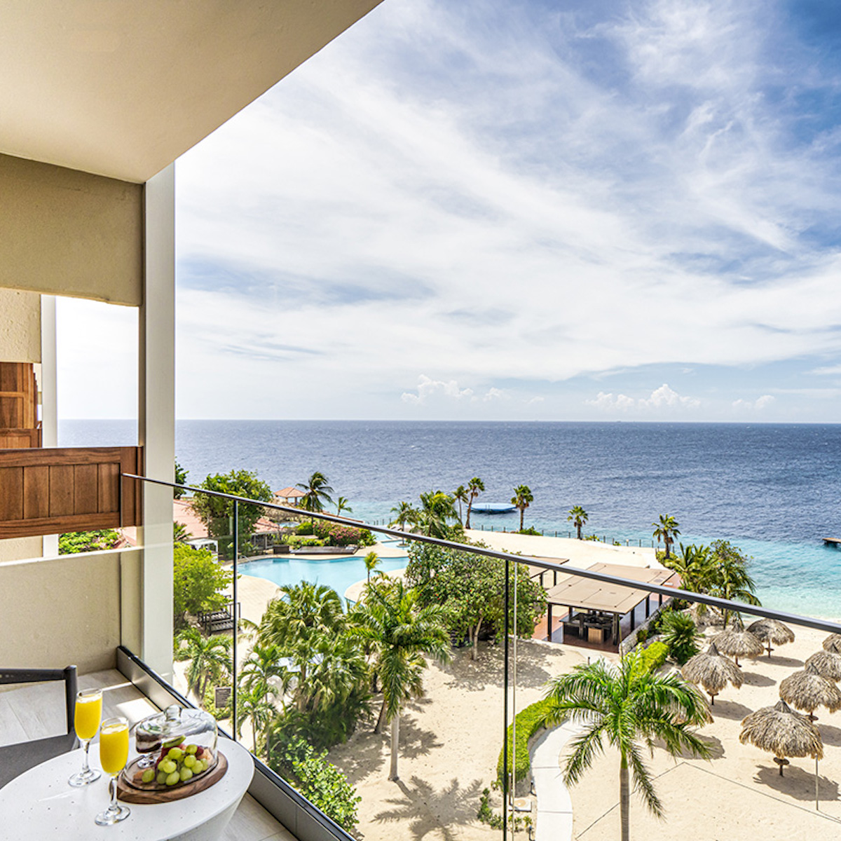 View from a balcony showing a destination wedding at Dreams Curacao with a pool, palm trees, and the ocean under a clear blue sky, with a table featuring breakfast items.