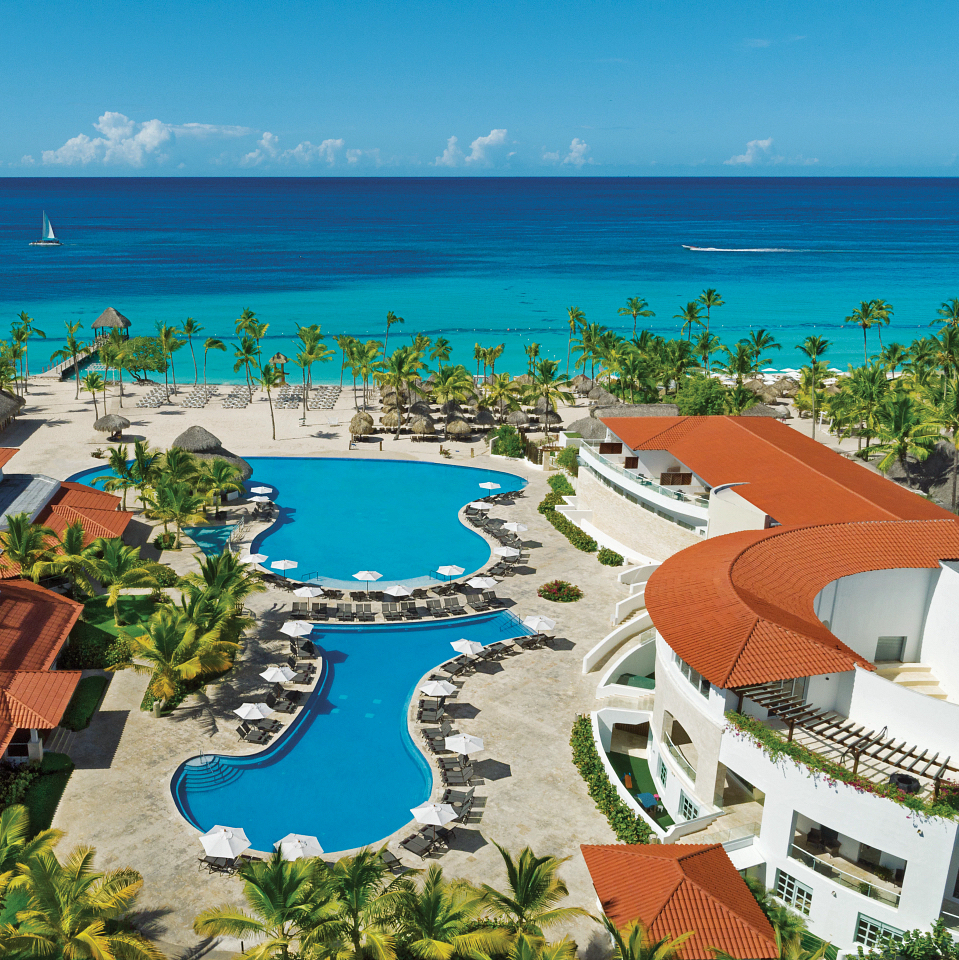 Aerial view of Dreams Dominicus, perfect for a destination wedding, with a large swimming pool, red-tiled villas, palm trees, and a clear view of the blue ocean.