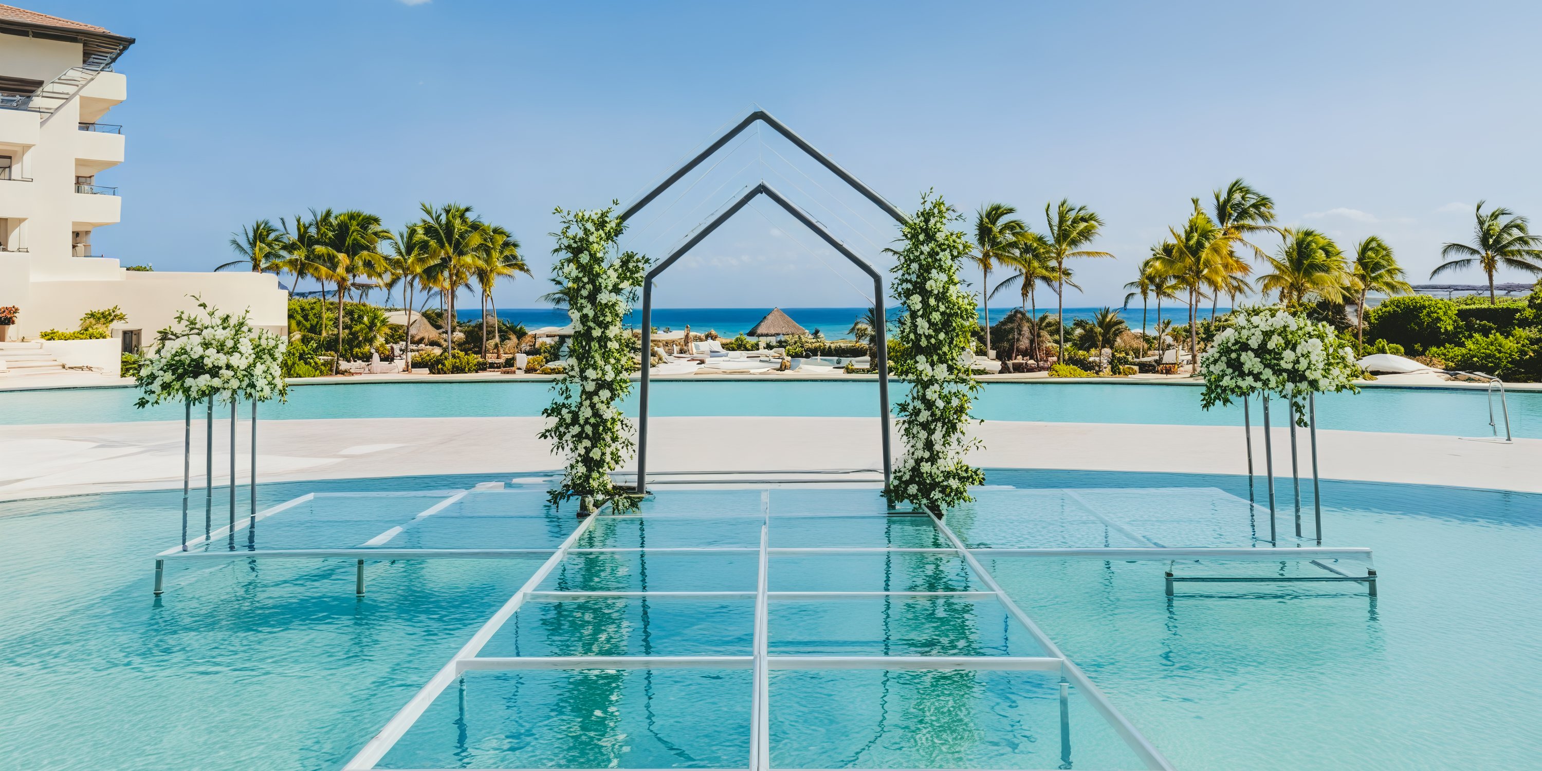A transparent walkway gracefully stretches over the pool, adorned with a floral archway, surrounded by palm trees and offering a stunning ocean backdrop—an idyllic setting for a destination wedding.