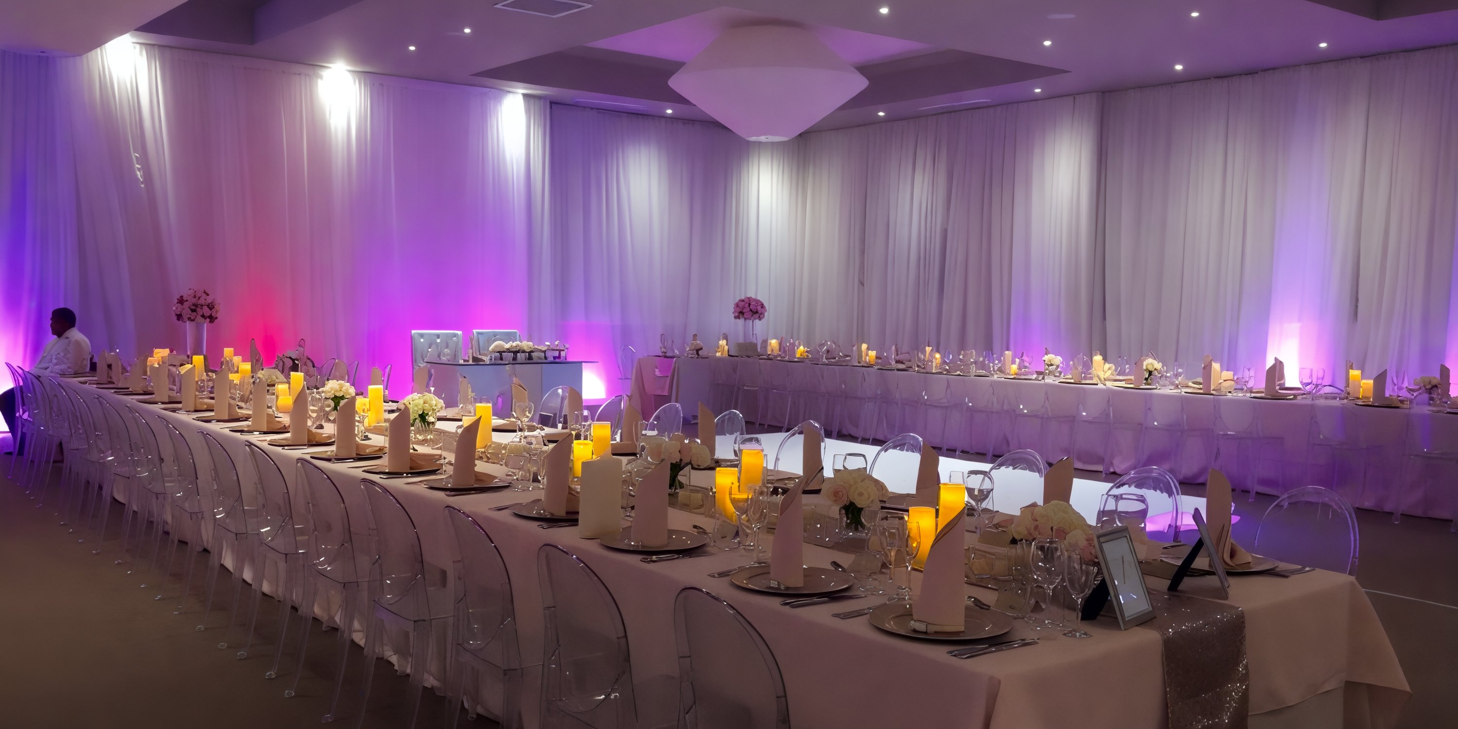 Experience a destination wedding with an Elegant Dreams Onyx setup, featuring long tables adorned with clear chairs and bathed in soft purple lighting.