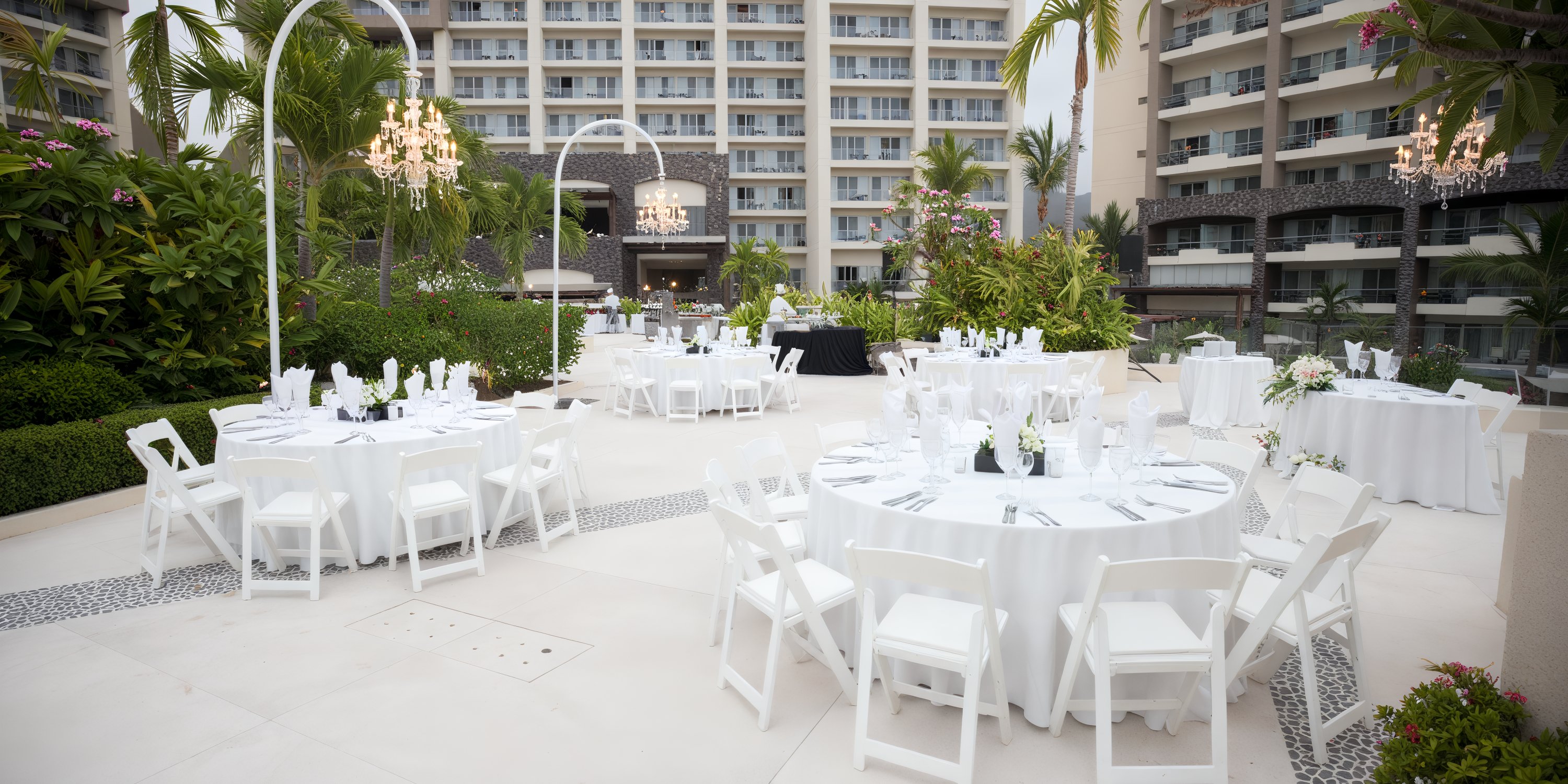 Setup for an outdoor destination wedding features round tables, elegant white chairs, and a picturesque building in the background.