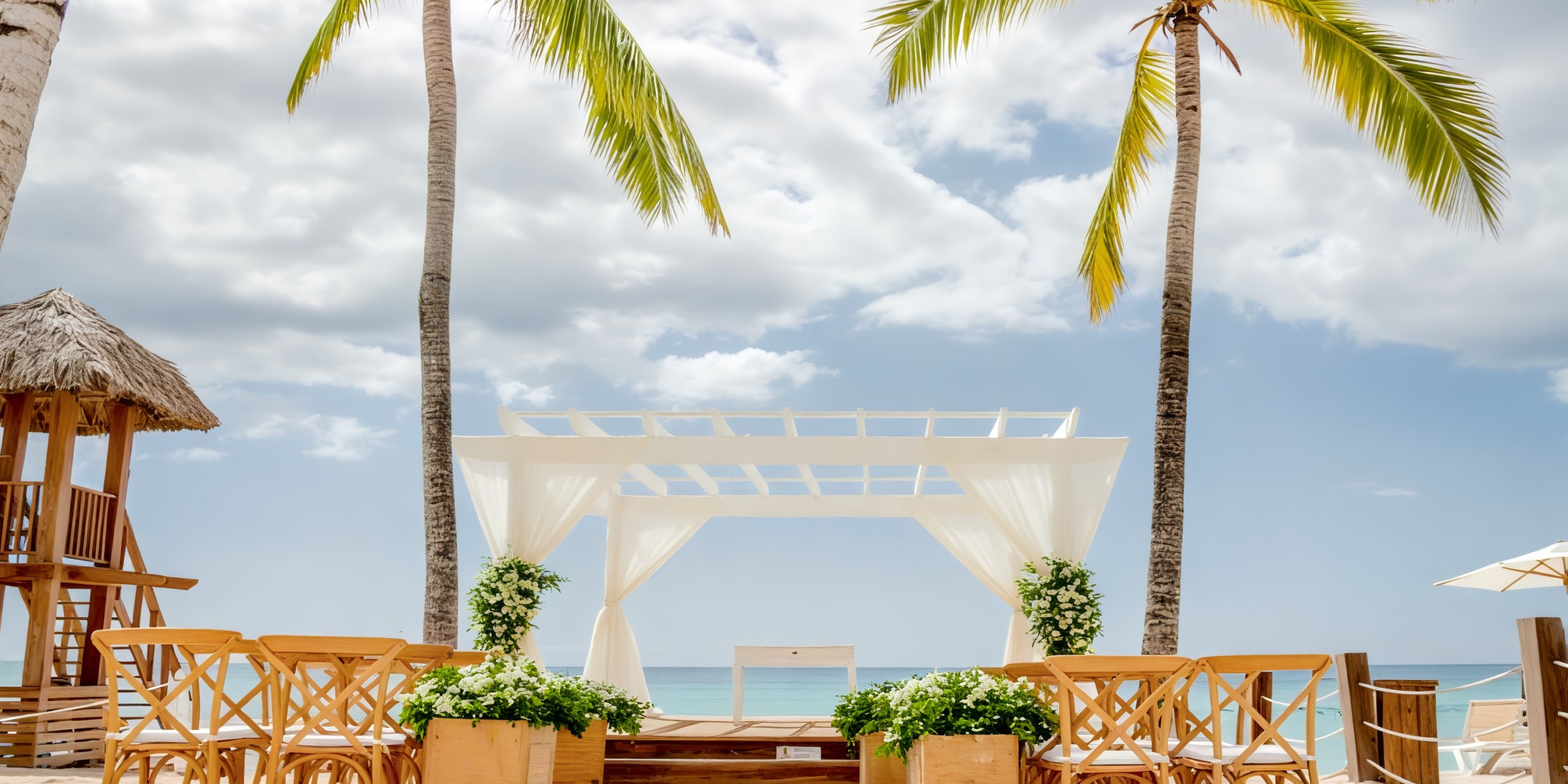 Dreams Dominicus offers a breathtaking destination wedding setup, featuring wooden chairs beneath a pristine white canopy, surrounded by palm trees with a stunning ocean view.