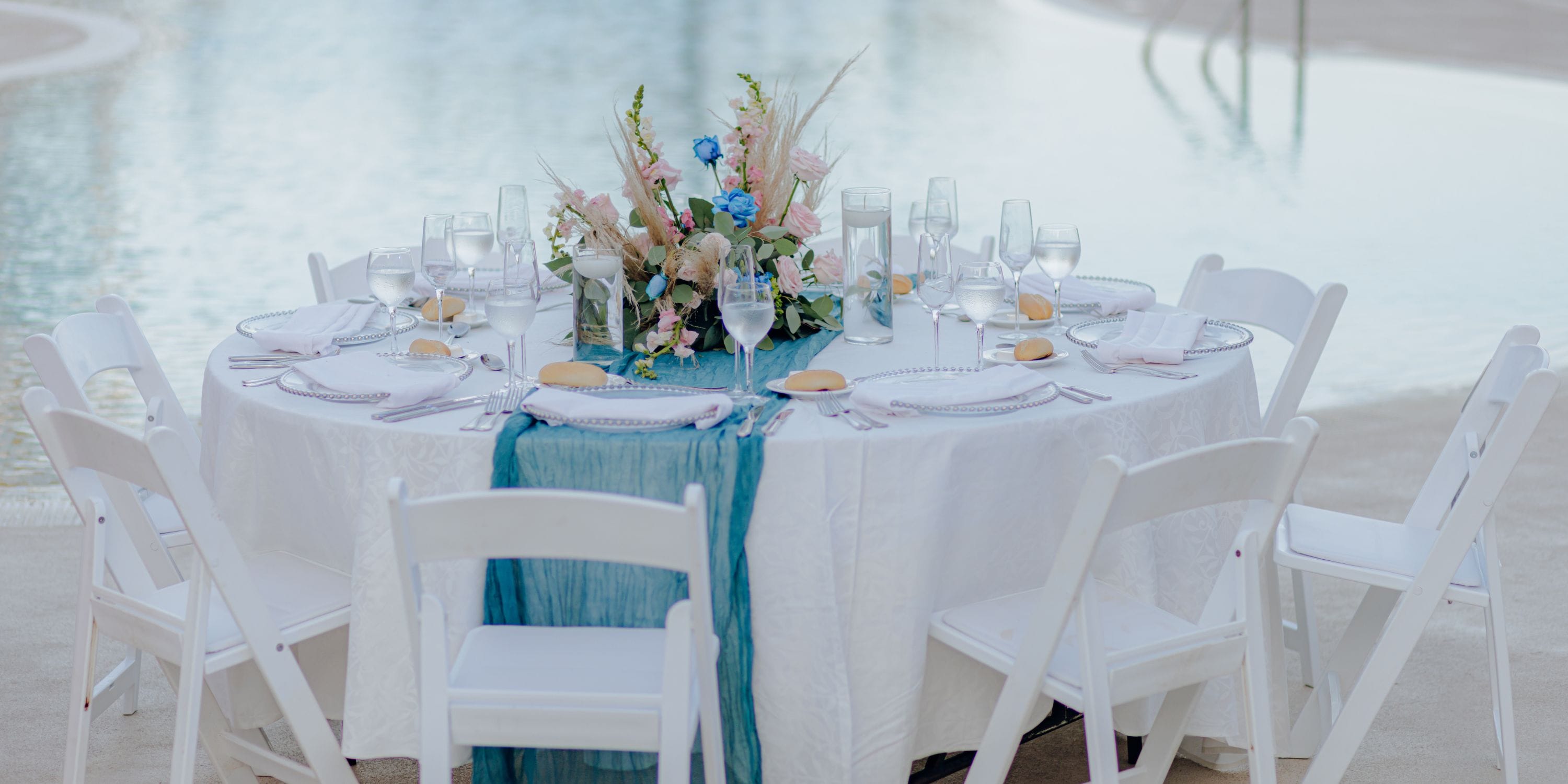 Round table set for a destination wedding by the pool at Dreams Jade Riviera Cancun with white chairs and floral centerpiece.