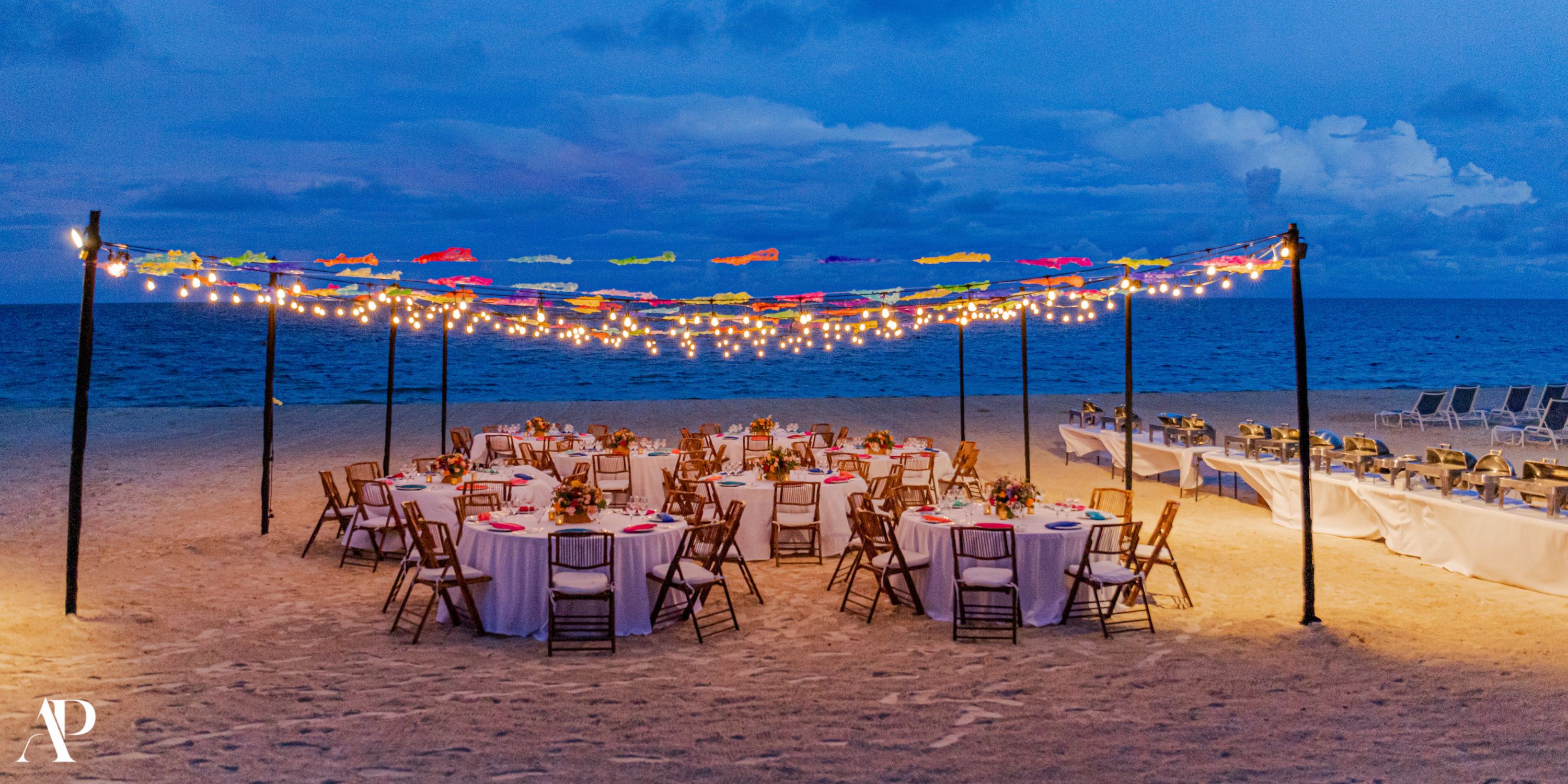 Round tables, chairs, and string lights set for a destination wedding on the sandy beach at dusk.