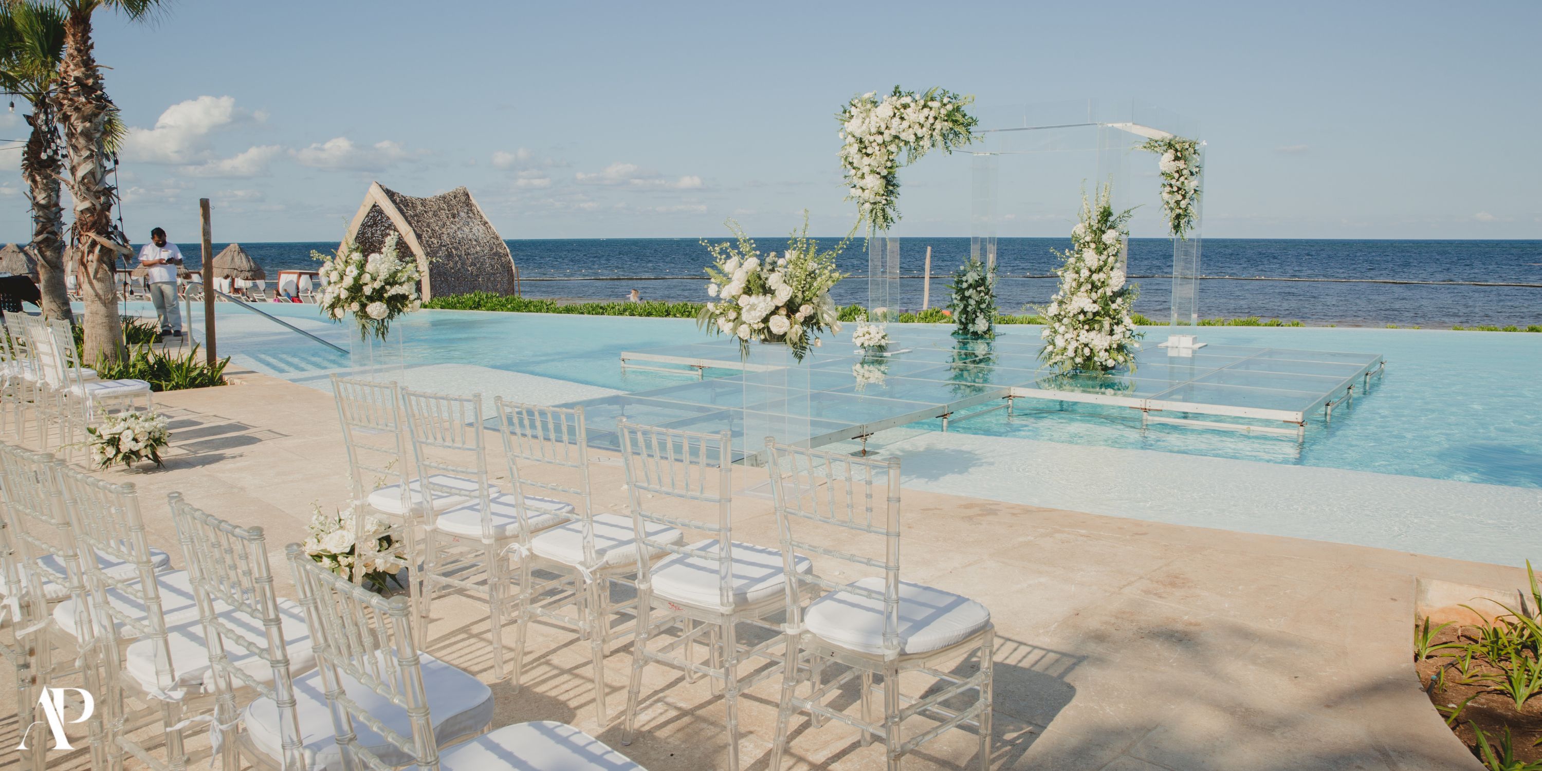 White chairs are arranged for a destination wedding facing a floral glass altar over a pool by the ocean.
