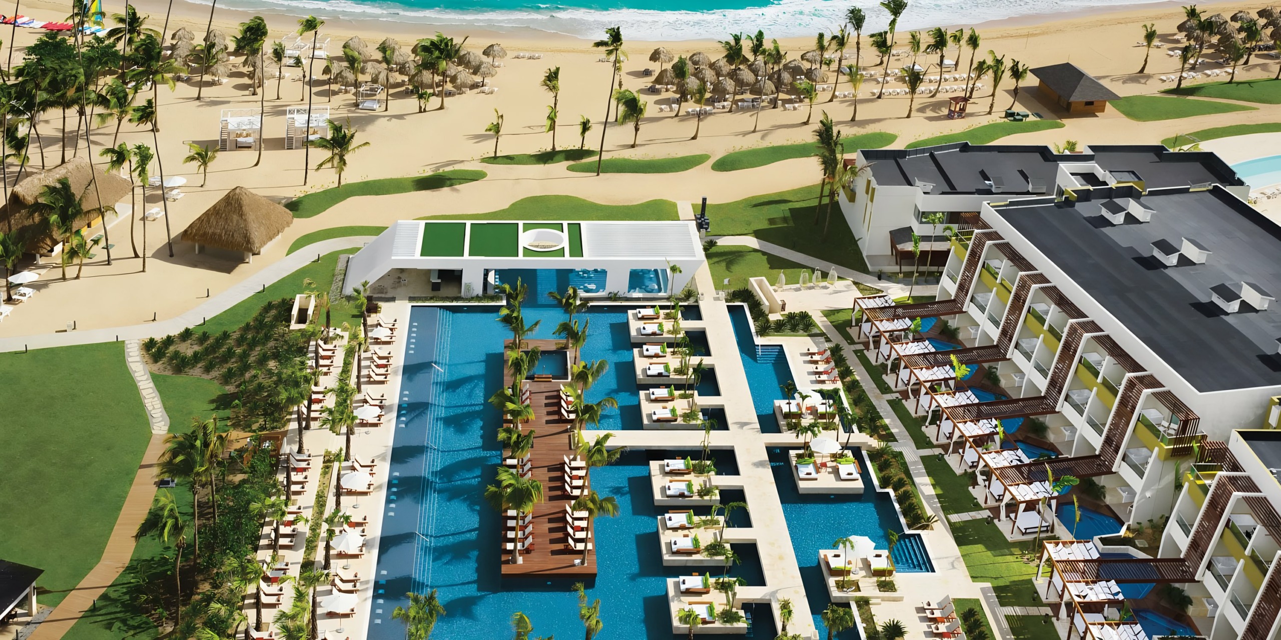 Aerial view of Dreams Onyx, a luxury resort with a large pool area, surrounded by buildings and palm trees, adjacent to a sandy beach.