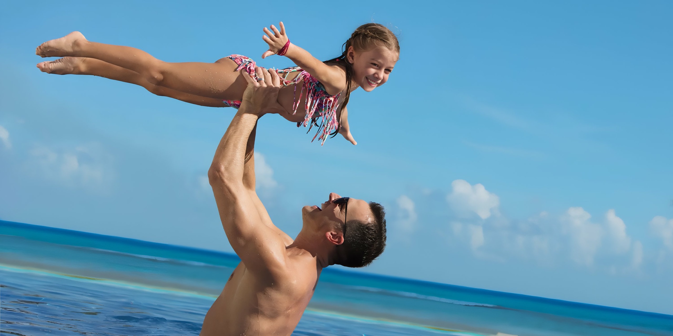 A man joyfully lifting a young girl above his head at a sunny beach with a clear blue sky and ocean in the background wearing Dreams Onyx.