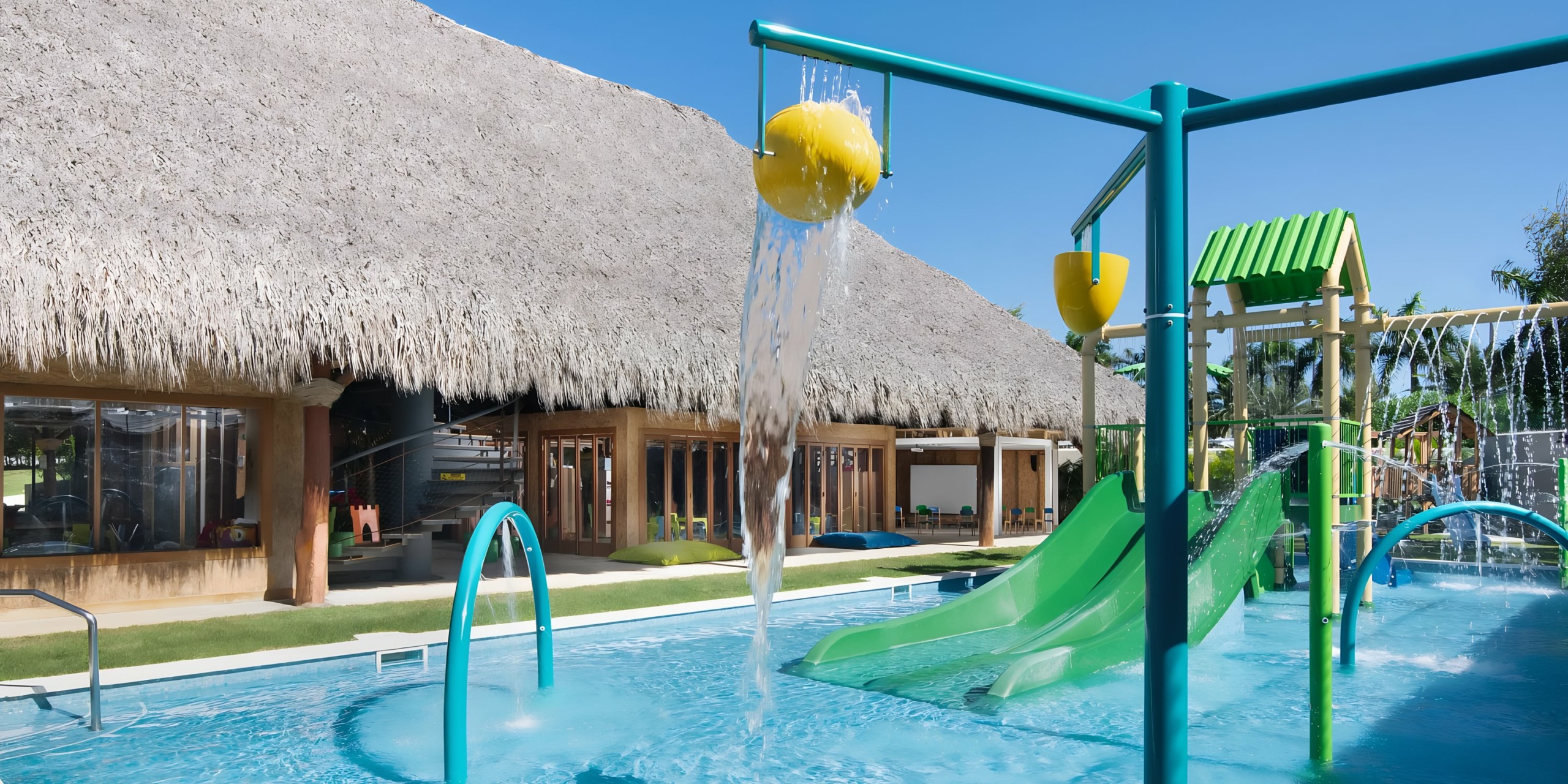 Dreams Onyx playground with slides and splash features in a pool, adjacent to a building with a large thatched roof on a sunny day.