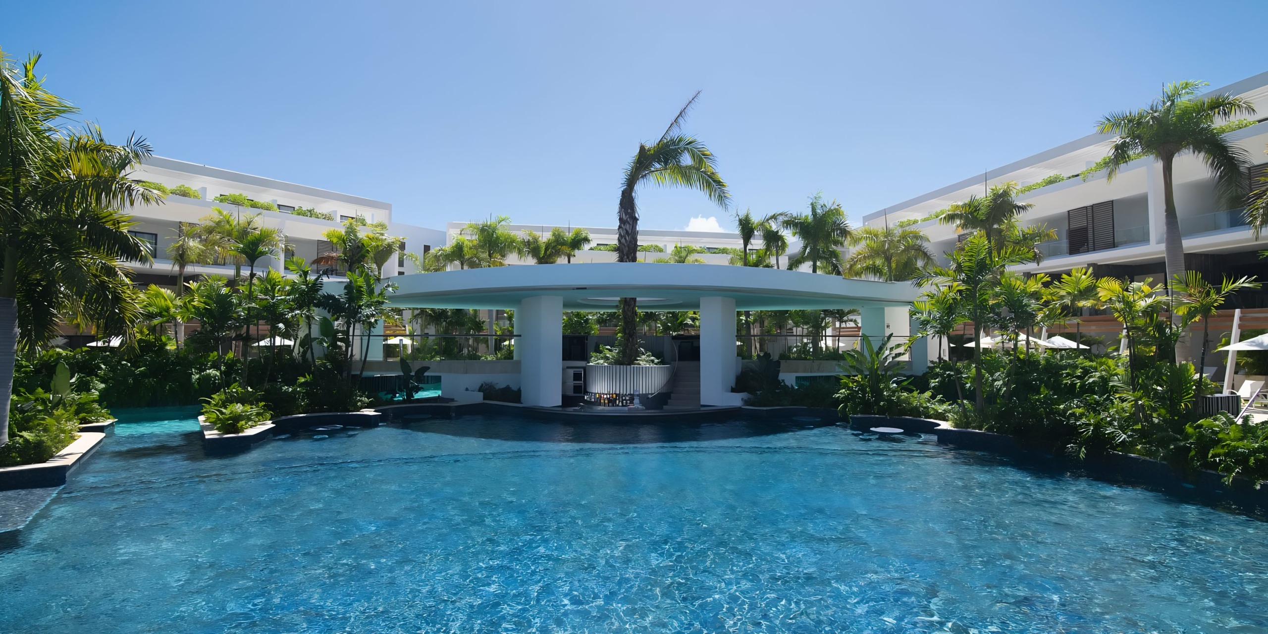 Modern Dreams Onyx resort pool surrounded by lush greenery, with a central bar under a white pavilion and flanked by symmetrical white buildings.