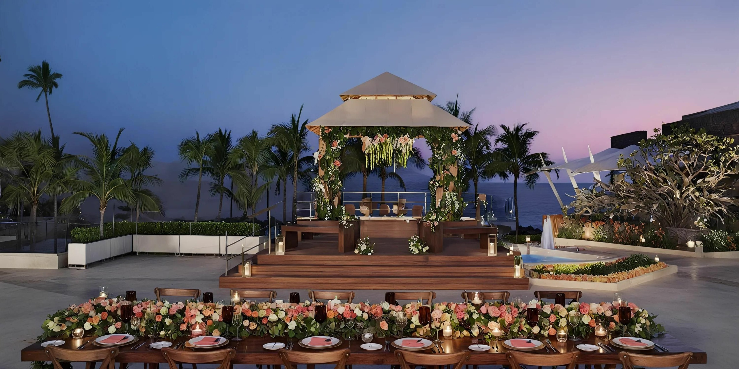 A beachside event setup at Dreams Vallarta Bay features a wooden stage adorned with flowers, surrounded by palm trees. A long dining table with flower centerpieces, plates, and cutlery is in the foreground under the twilight sky above.