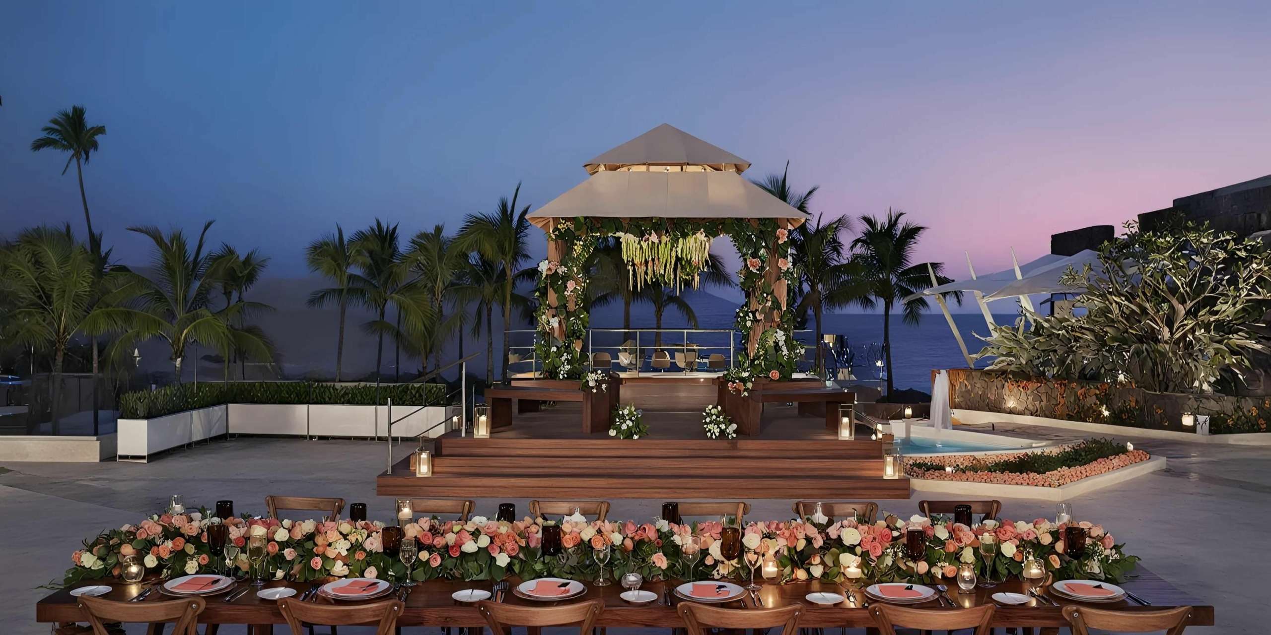 An outdoor wedding setup with a decorated altar on a platform overlooking the ocean at sunset. The dining table is adorned with flowers and candles, and palm trees are in the background.