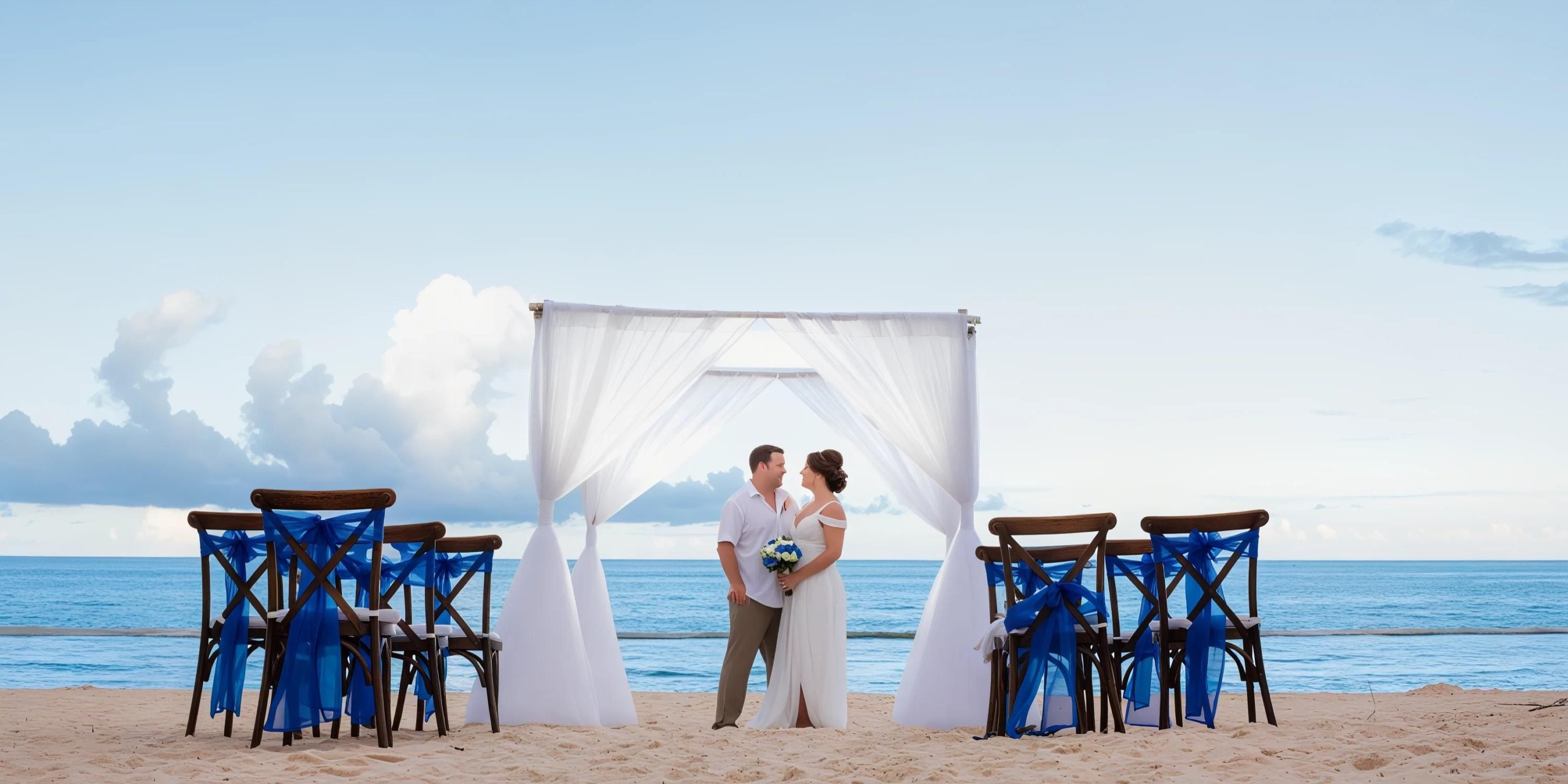 At Hyatt Zilara Cap Cana, a couple stands under a pristine white archway on the sandy beach, with the tranquil ocean and expansive sky creating a stunning backdrop. Empty chairs adorned with elegant blue sashes are neatly arranged on both sides.
