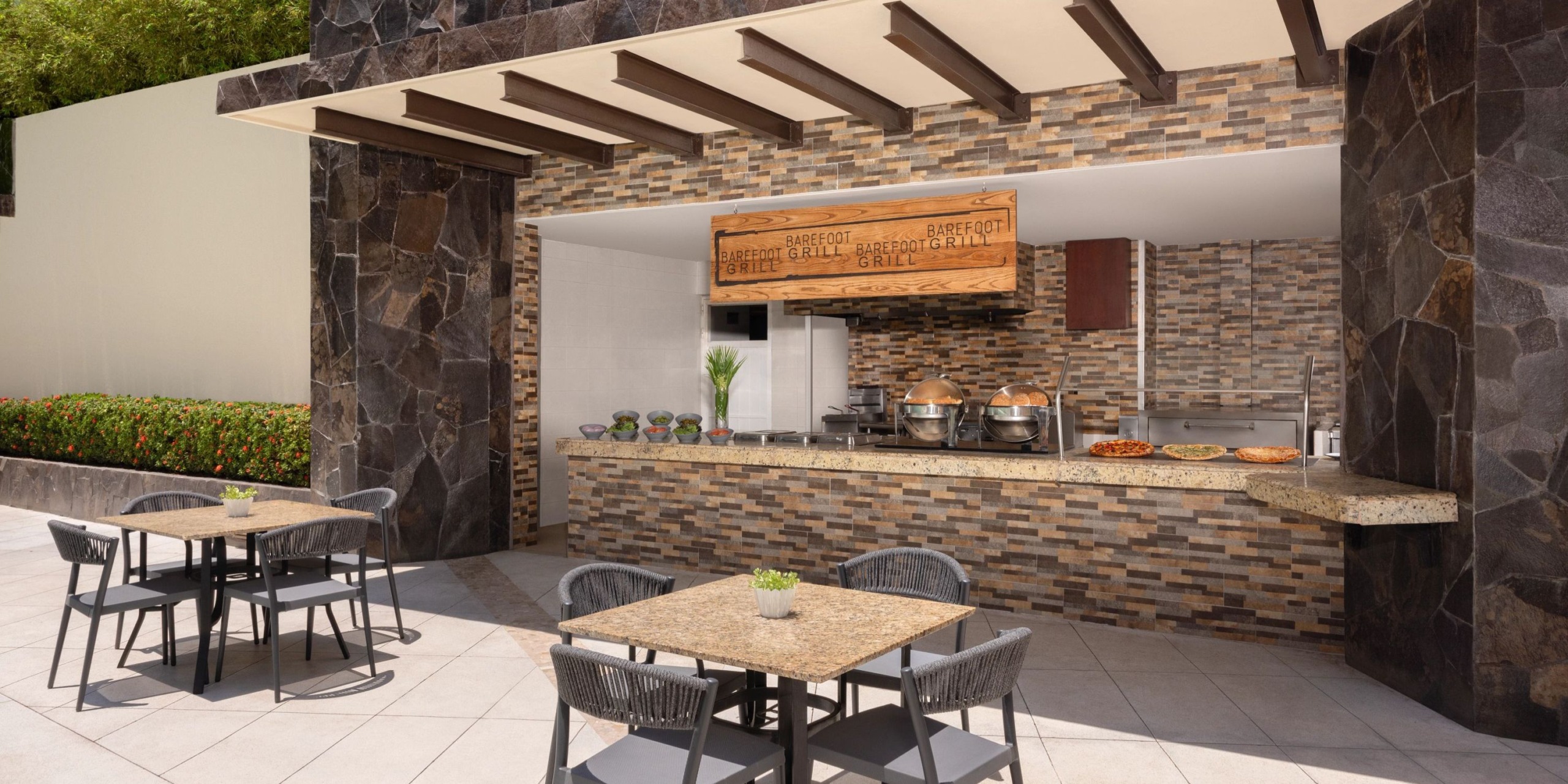 An outdoor setting at Dreams Vallarta Bay showcases a stone design, wooden beams, and patio seating, featuring a buffet counter with an array of dishes.