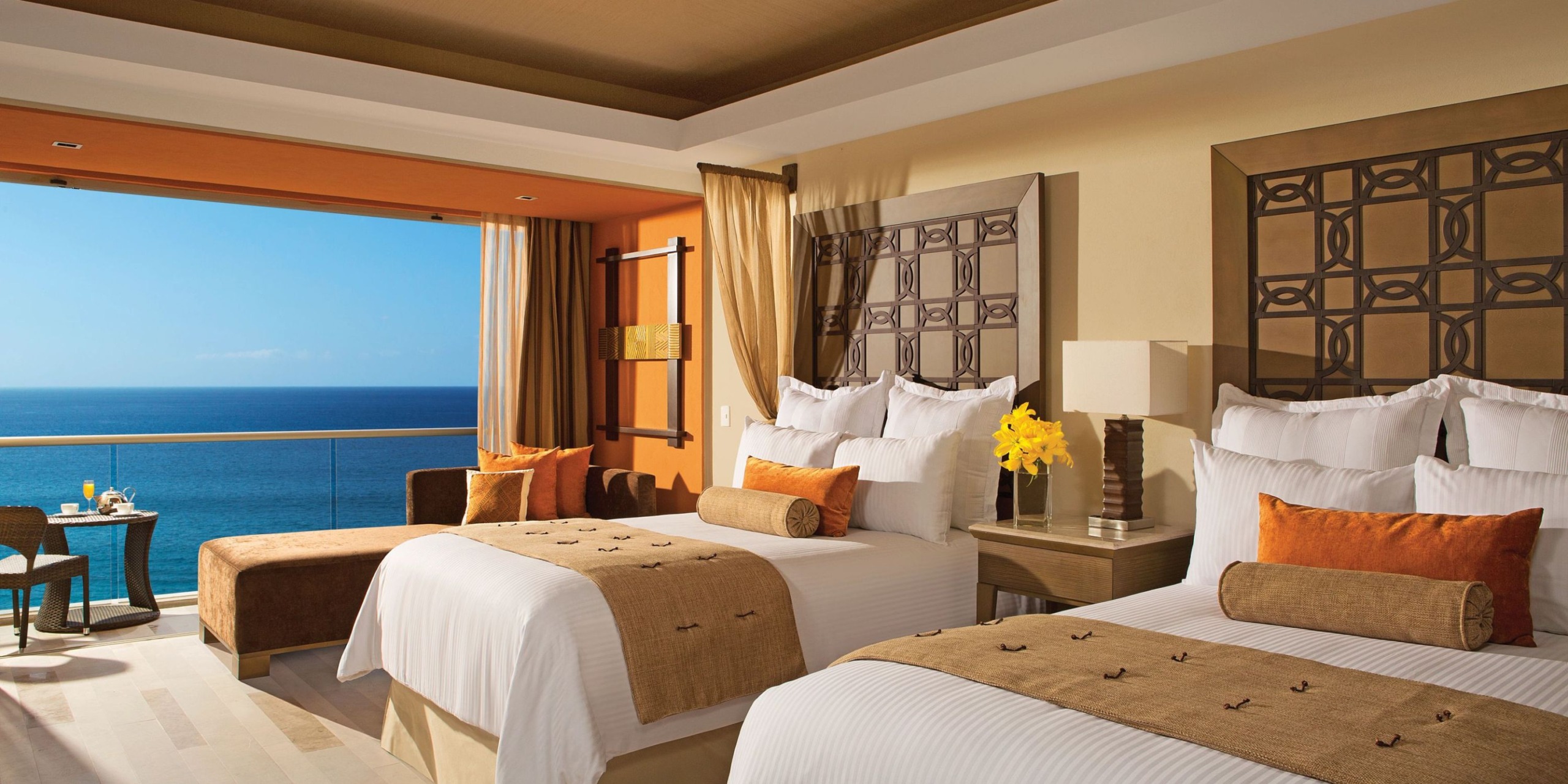 Stay at Dreams Vallarta Bay in a hotel room featuring two double beds, a cozy small table with chairs, a comfortable sofa, and a balcony with breathtaking ocean views. The decor showcases elegant beige tones complemented by decorative pillows.