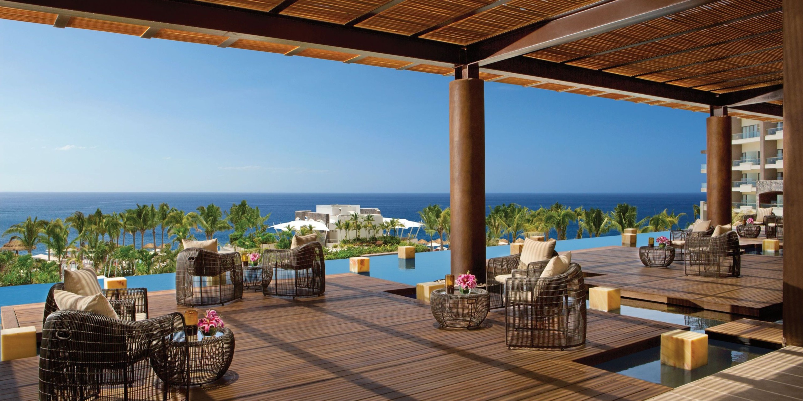 Experience the luxurious patio of Dreams Vallarta Bay, featuring elegant wicker chairs and tables that overlook a sparkling pool, majestic palm trees, and the serene ocean, all under a beautifully crafted wooden pergola.