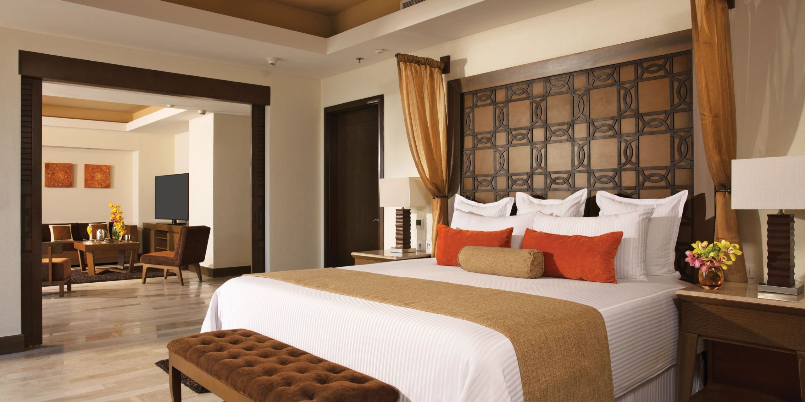 Experience the opulence of the Dreams Vallarta Bay suite, featuring a spacious bed embellished with orange and white cushions, complemented by a wooden headboard and coordinating nightstands. Nearby is a comfortable seating area complete with a table and chairs, alongside a wall-mounted TV for your entertainment.